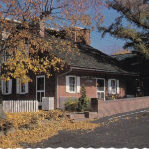 A small brick building with a wood-shingled roof, resembling the Gettysburg Postcard Jennie Wade House, sits behind a white picket fence and autumn trees. A sign above the door reads "Hessian House.