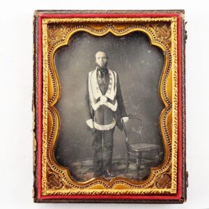 A Masonic Daguerreotype featuring a man in formal attire with a sash, standing beside a chair, presented in an ornate gold and red case.