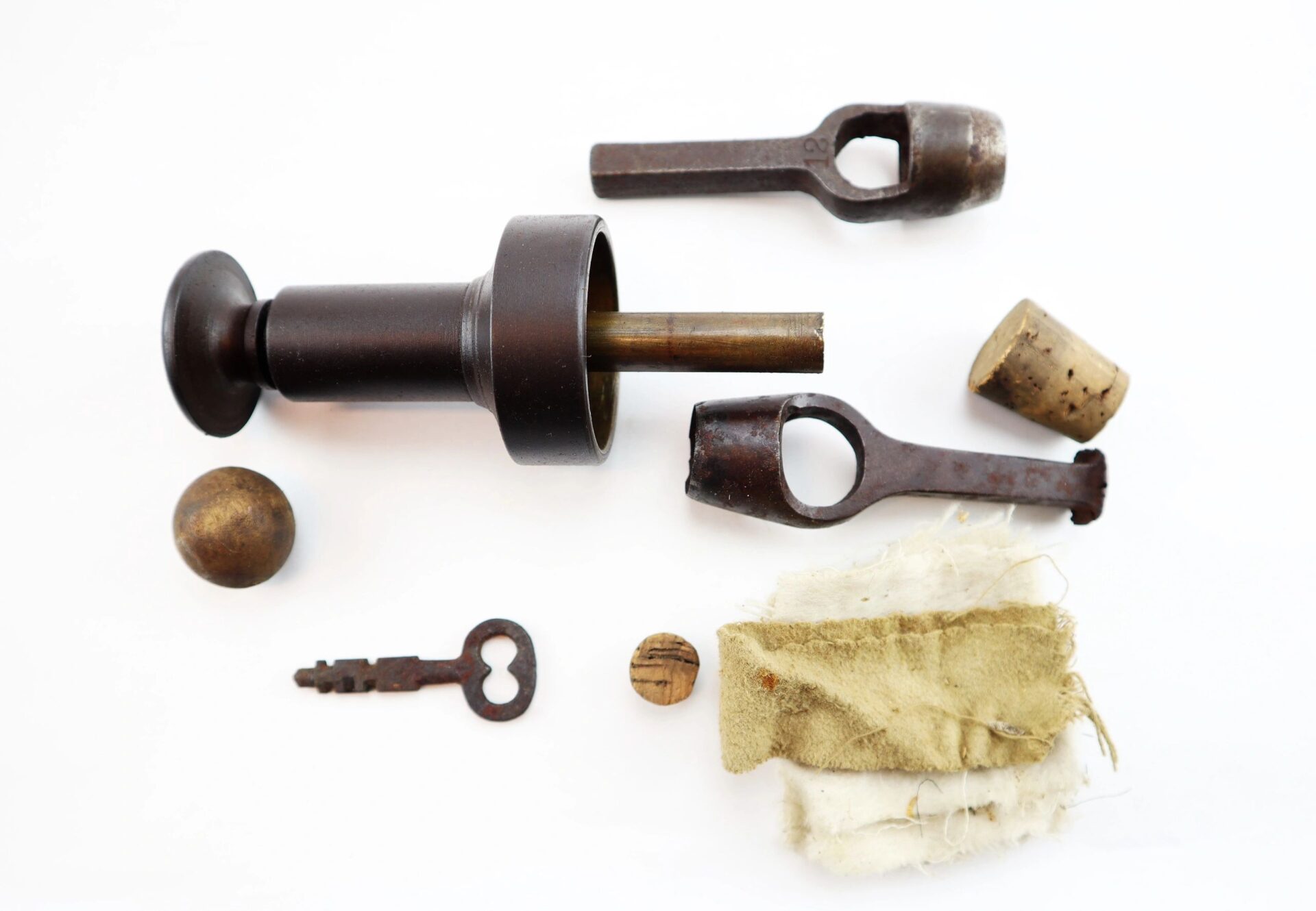 A collection of vintage objects from the "Target Rifle" Box Sharpshooters Display, arranged on a white surface—including a syringe, stoppers, key, wooden ball, wrench-like tools, button, and cloth.
