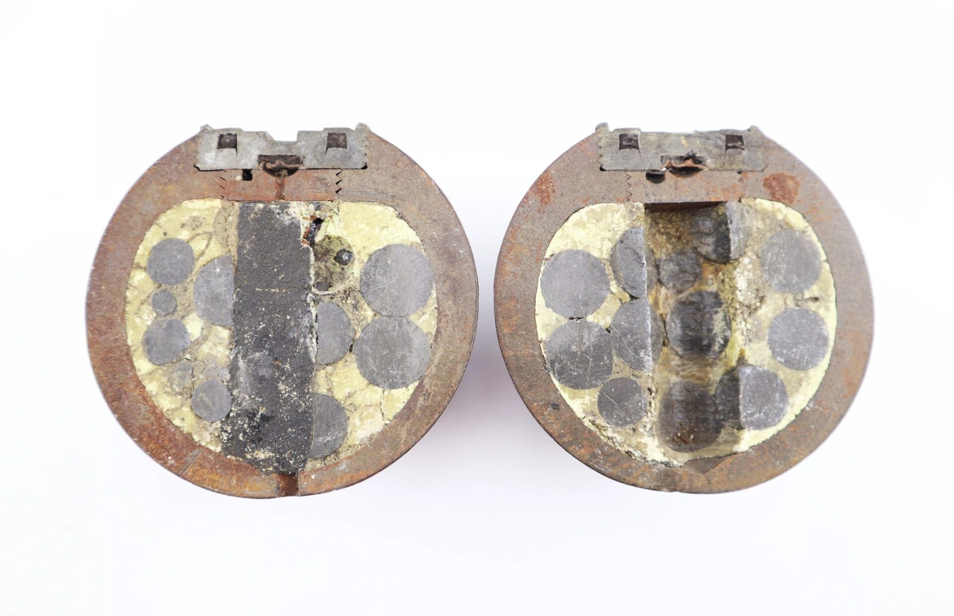 Two rusted, circular metallic objects with hinged lids—Confederate 6 Pound Borman Artillery Shells—are open, revealing interiors filled with black circular sections embedded in a yellowish material.