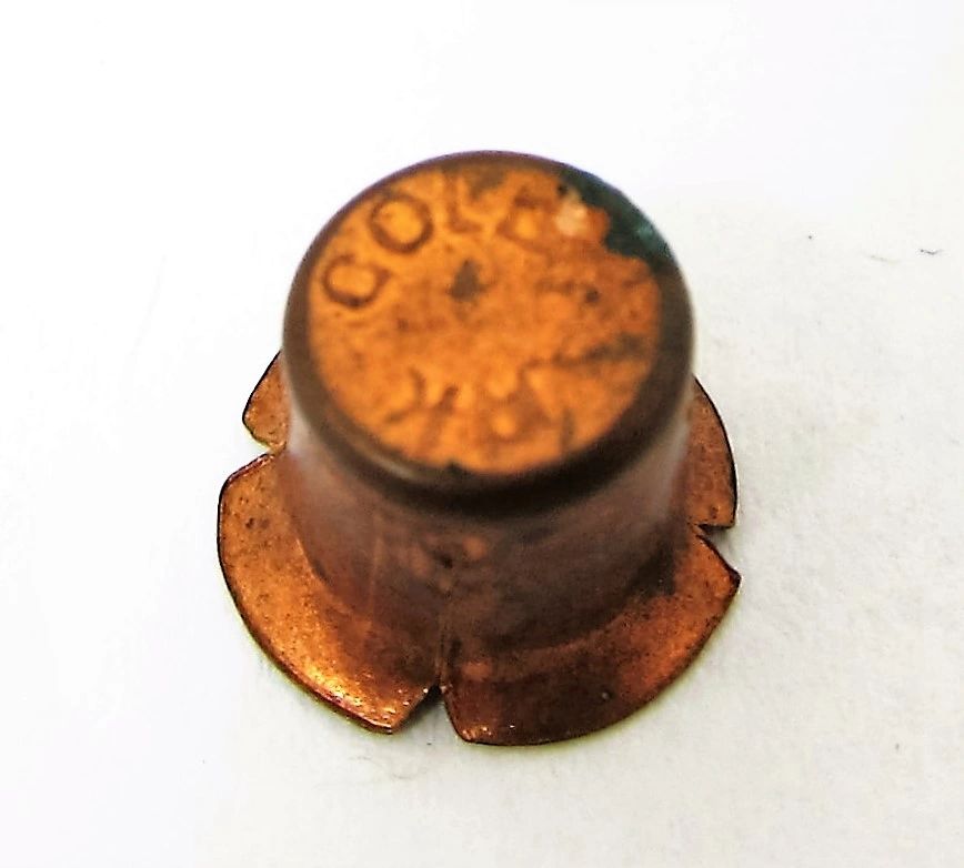 Close-up of a spent Percussion Cap with "COLT" and "K" stamped on top, viewed from above, with petal-like markings at the base.