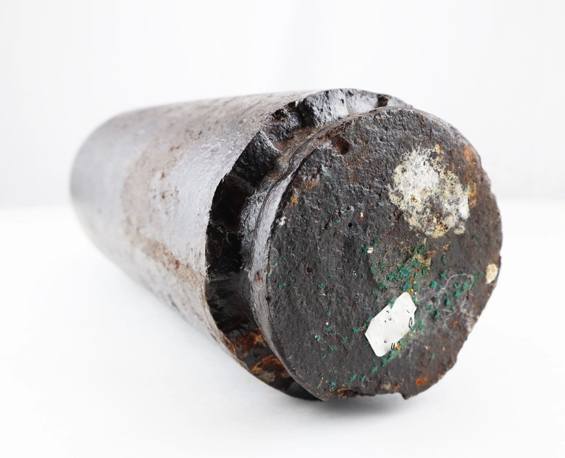 A US 4.2” 30-Pounder Parrott Common Shell, featuring a rusted cylindrical metal body with chipped edges and green and white paint marks on one end, is displayed lying on a white surface.