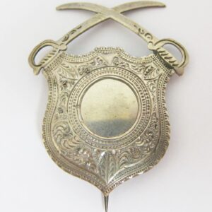 The Cavalry Corps Badge is a silver ornamental emblem featuring a detailed shield with intricate engravings and two crossed swords at the top.