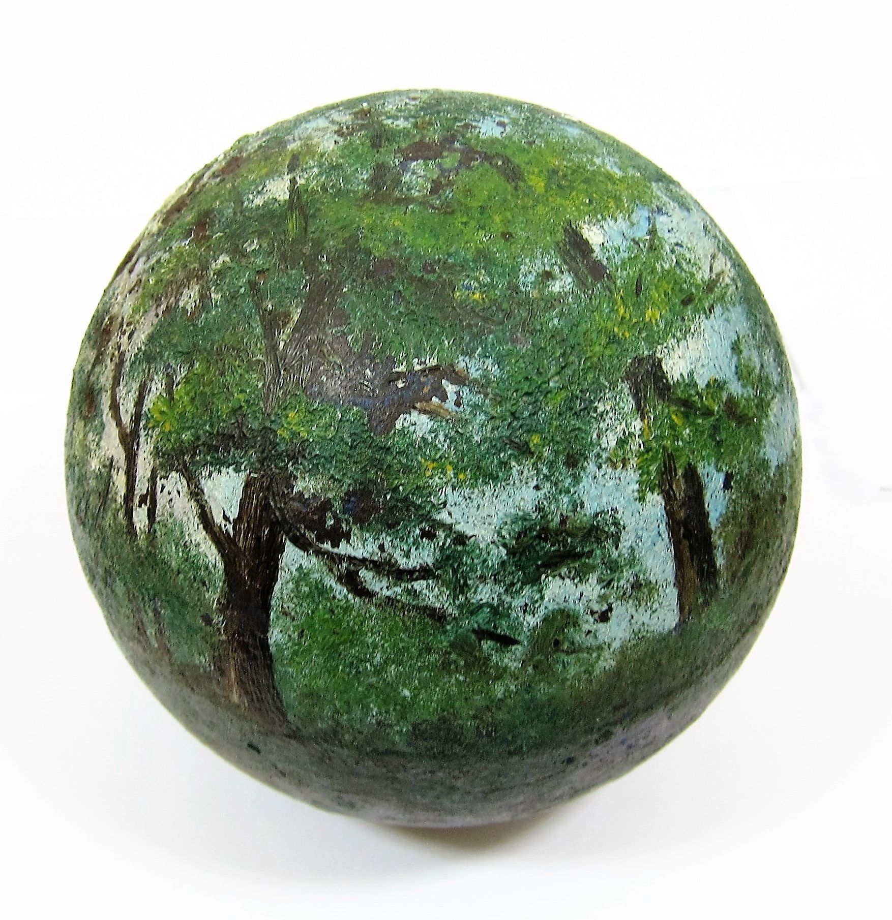 The Painted Cannon Ball is a round object featuring a landscape of green trees and foliage painted on its surface against a white background.