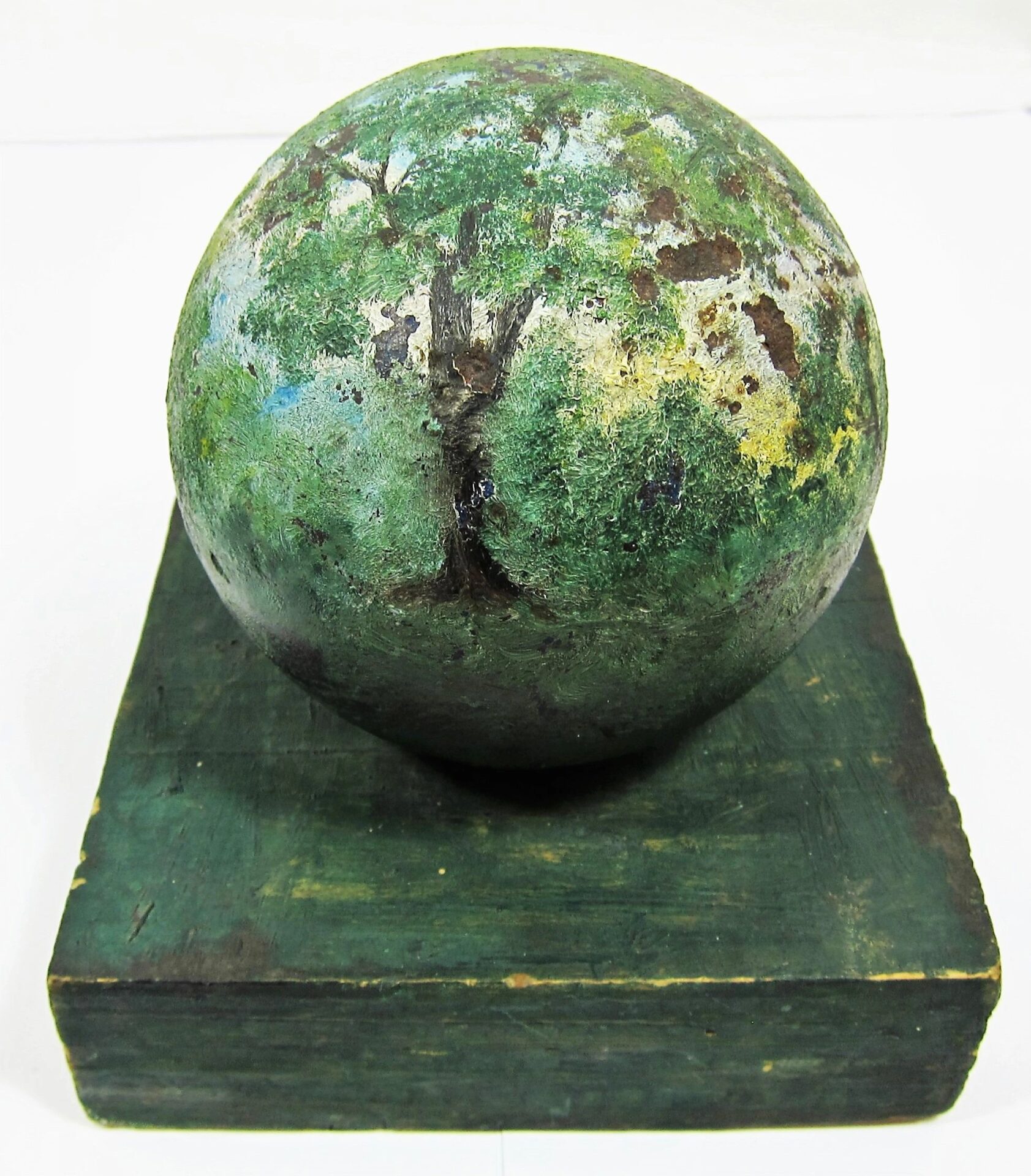 A Painted Cannon Ball, green with weathered tree-like patterns, sits atop a matching square base against a plain background.