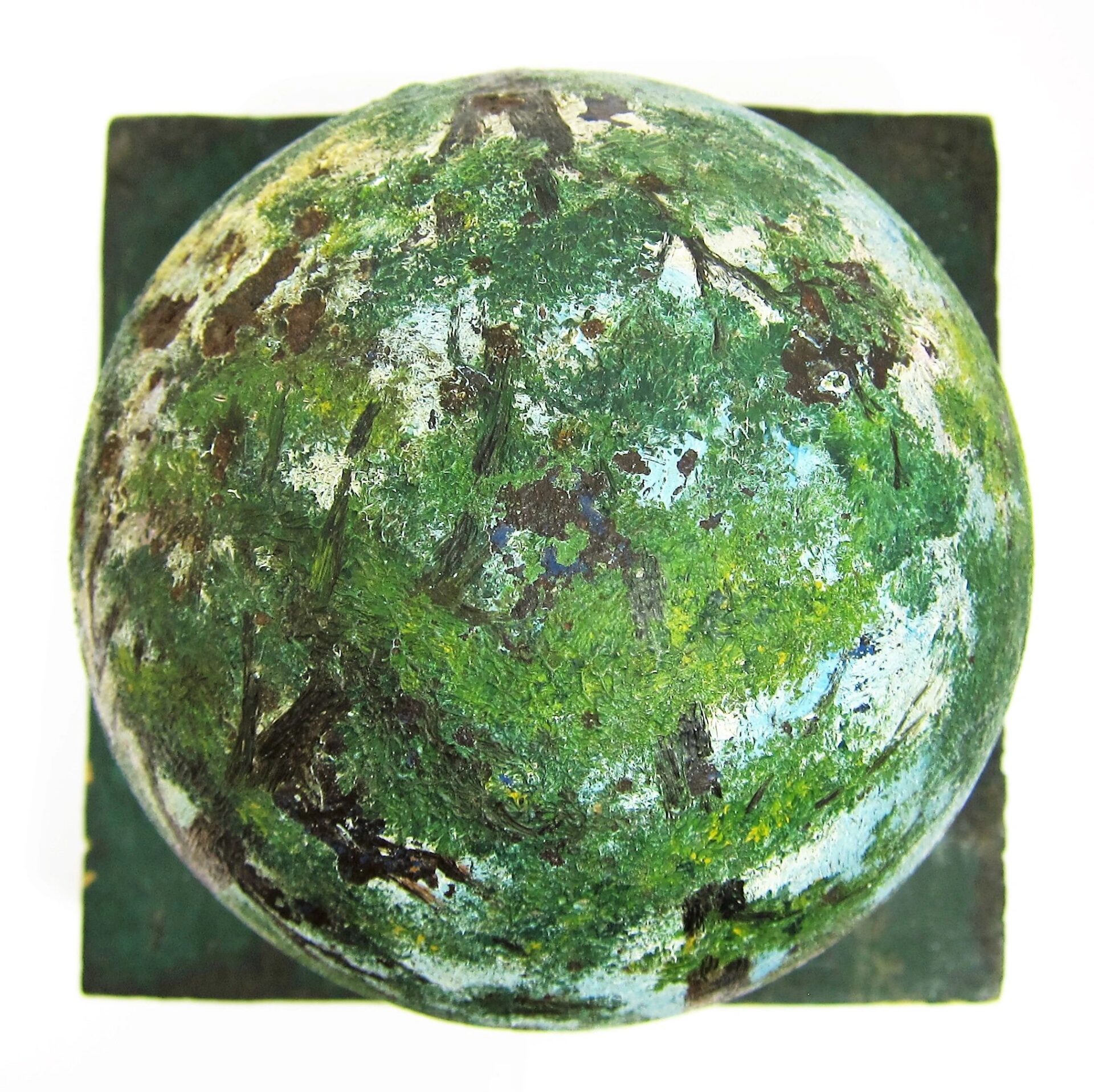 A Painted Cannon Ball with a mottled green and white textured surface sits centered on a square base, viewed from above.