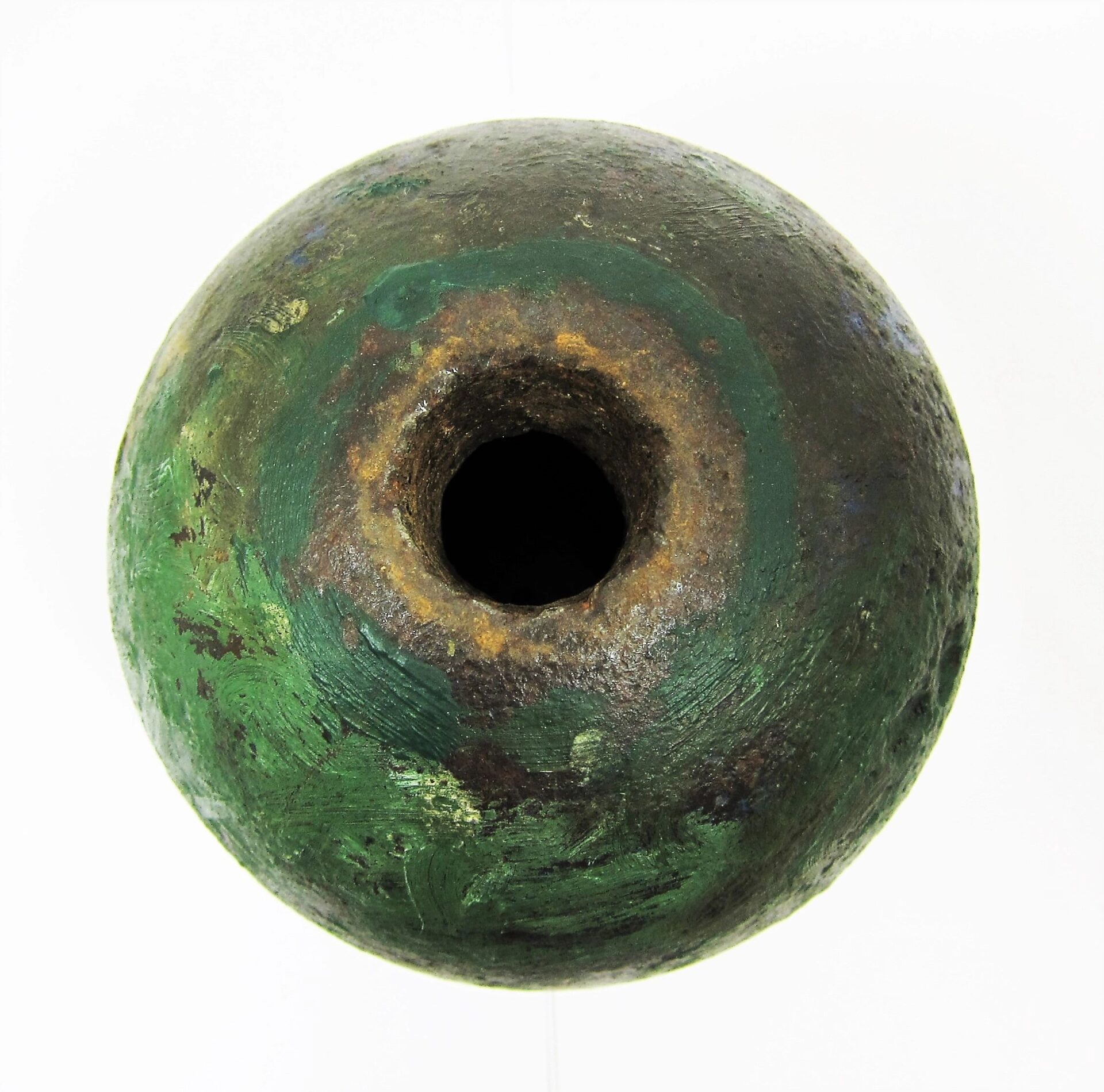 A painted cannon ball with a large central hole and greenish corrosion is shown from above against a white background.