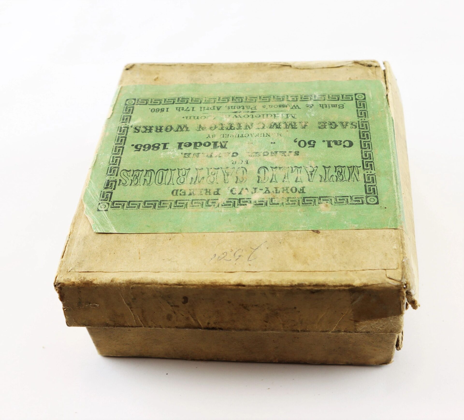 A worn, rectangular cardboard box labeled "Complete Box of 1865 Spencer Cartridges," featuring a green vintage label with text and decorative borders, is viewed at an angle.