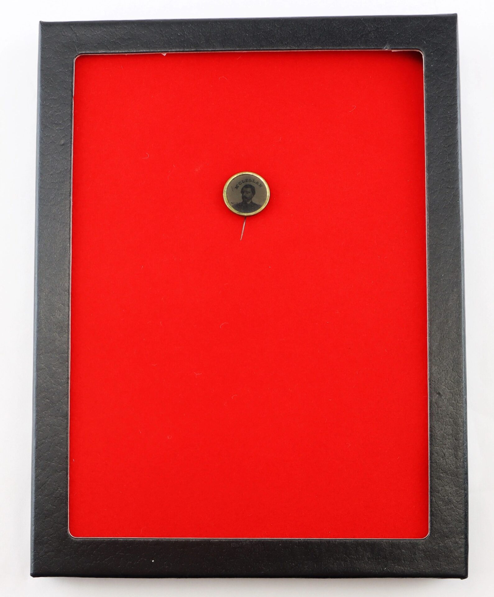 The George McClellan Ferrotype Pin, a small round pin with a portrait, is centered on a bright red background and framed in a black rectangular display case.