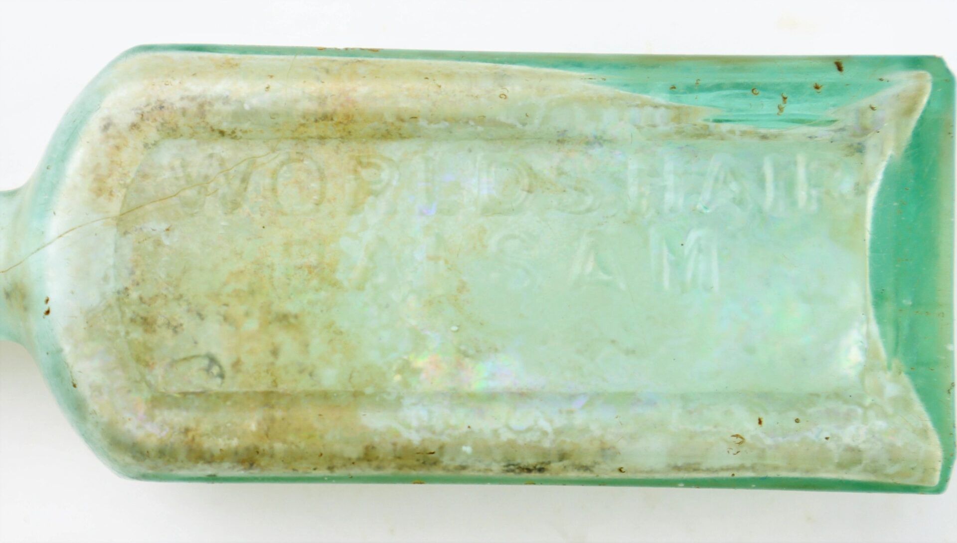 Close-up of an old, translucent Mrs. S.A. Adams Worlds Hair Balsam bottle from Brandy Station, Virginia, showing visible dirt and the partially readable embossed words "WORLD'S HAIR SAM" on its surface.