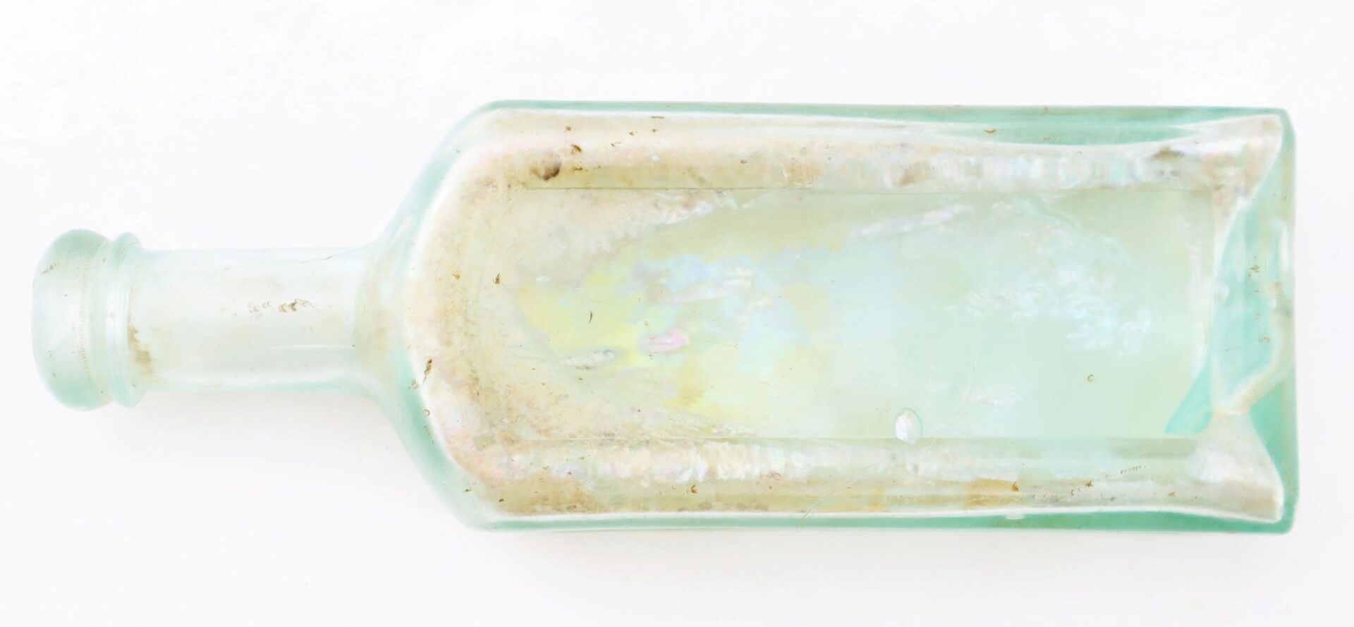 A rectangular, clear glass Mrs. S.A. Adams Worlds Hair Balsam Bottle from Brandy Station, Virginia, with a narrow neck and dirt and residue inside, is lying on its side against a white background.