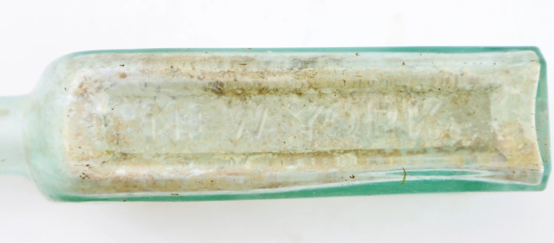 A close-up of the weathered Mrs. S.A. Adams Worlds Hair Balsam Bottle from Brandy Station, Virginia, an old rectangular greenish glass bottle with partially obscured "NEW YORK" engraved on its side.