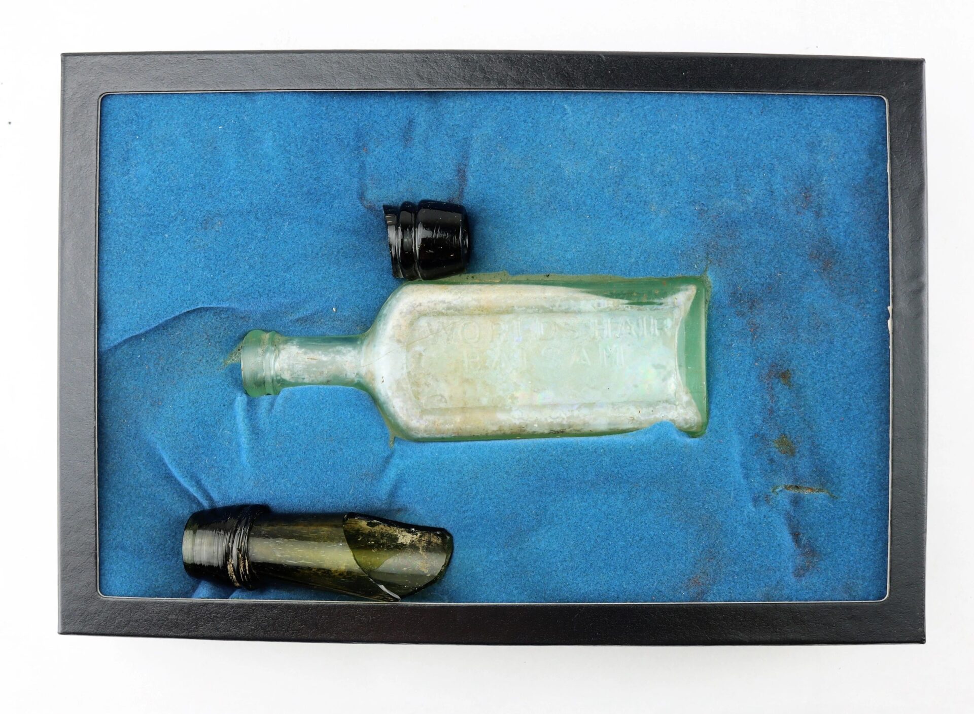 A display case with a blue background contains a Mrs. S.A. Adams Worlds Hair Balsam Bottle from Brandy Station, Virginia—one intact and one broken with two black caps.