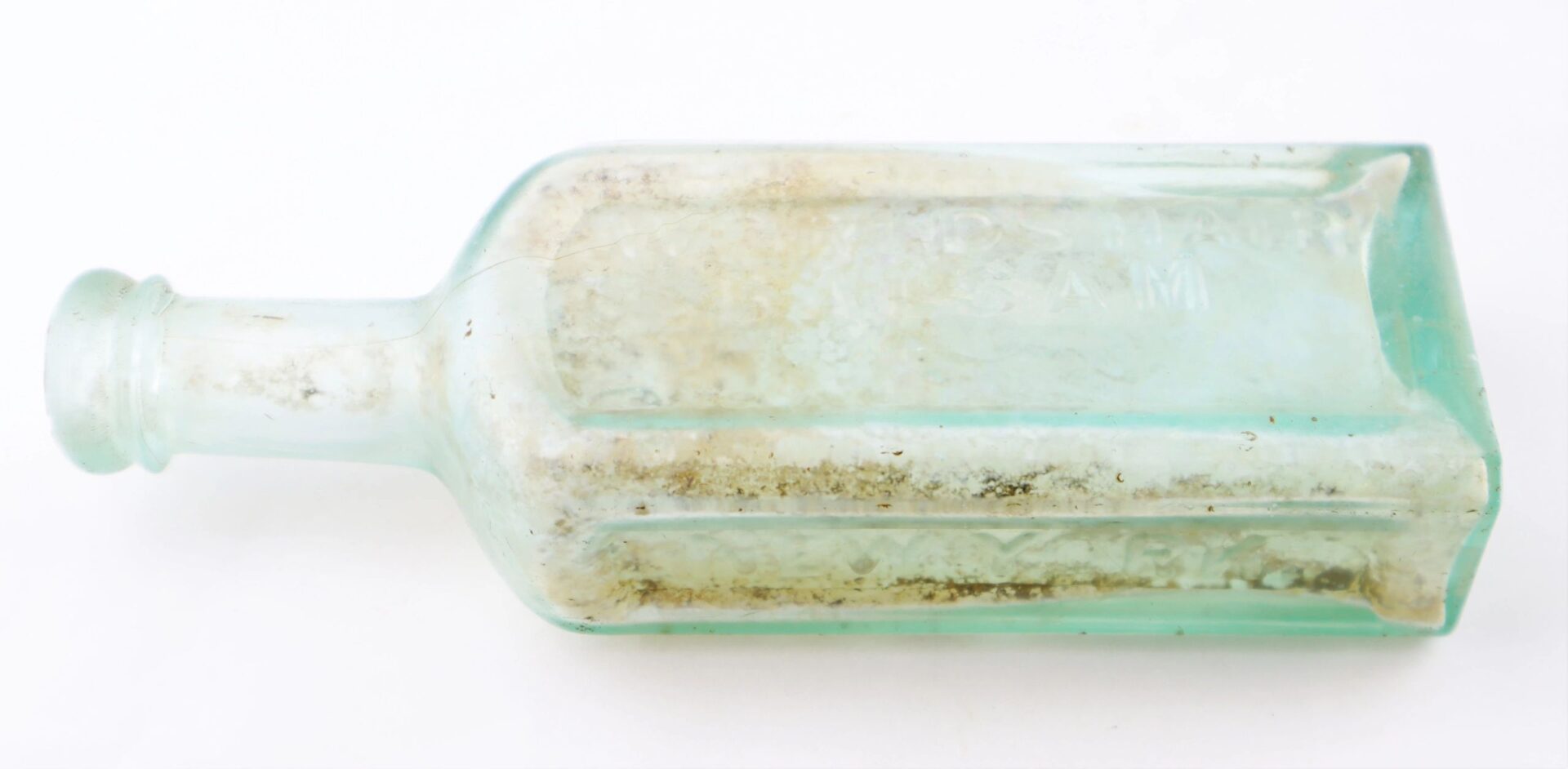 A clear, rectangular Mrs. S.A. Adams Worlds Hair Balsam Bottle from Brandy Station, Virginia, with visible dirt and residue inside, lies on its side against a white background.