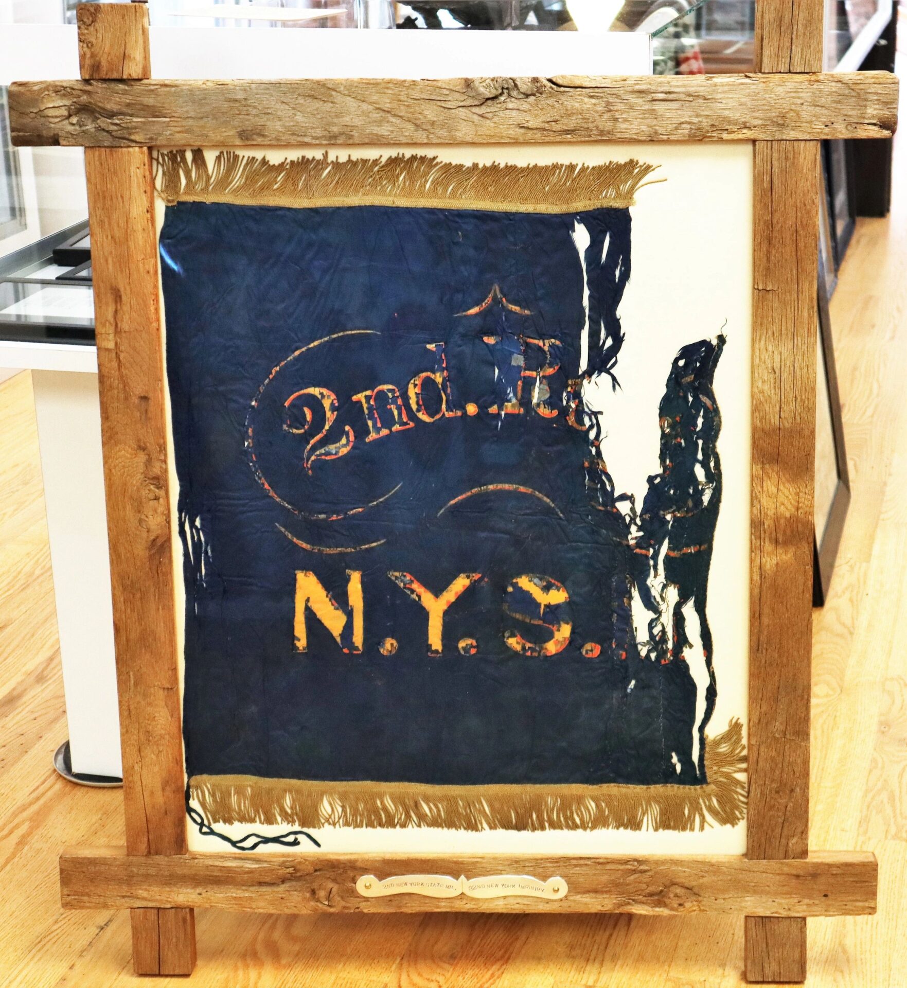 A tattered, framed blue Flank Marker Flag of the 2nd Regiment New York State Militia (82nd NY Infantry), Battle of Antietam and Gettysburg, with faded gold "2nd Lt. N.Y.S.," is displayed vertically on a museum wooden floor.