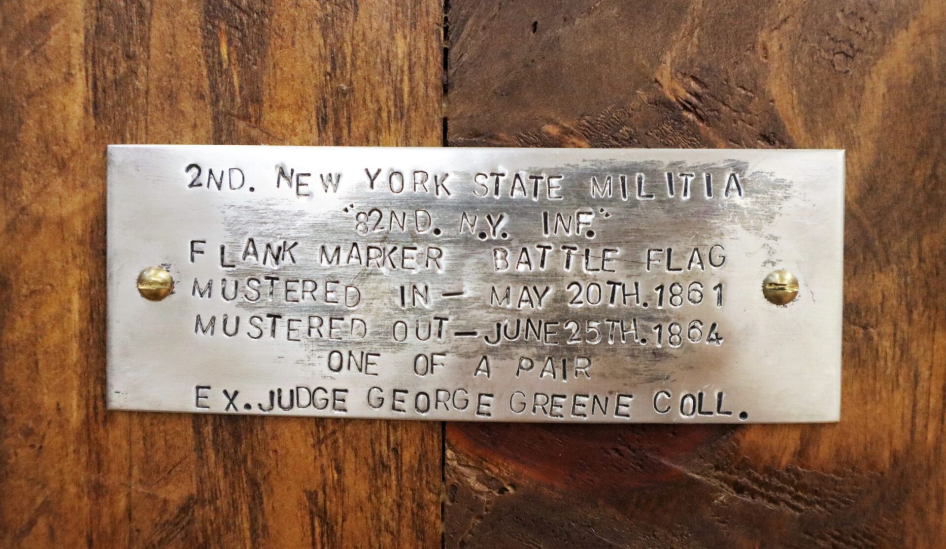 A metal plaque on wood with engraved text about the Flank Marker Flag, 2nd Regiment New York State Militia (82nd NY Infantry), referencing Antietam, Gettysburg, and the George Greene collection.
