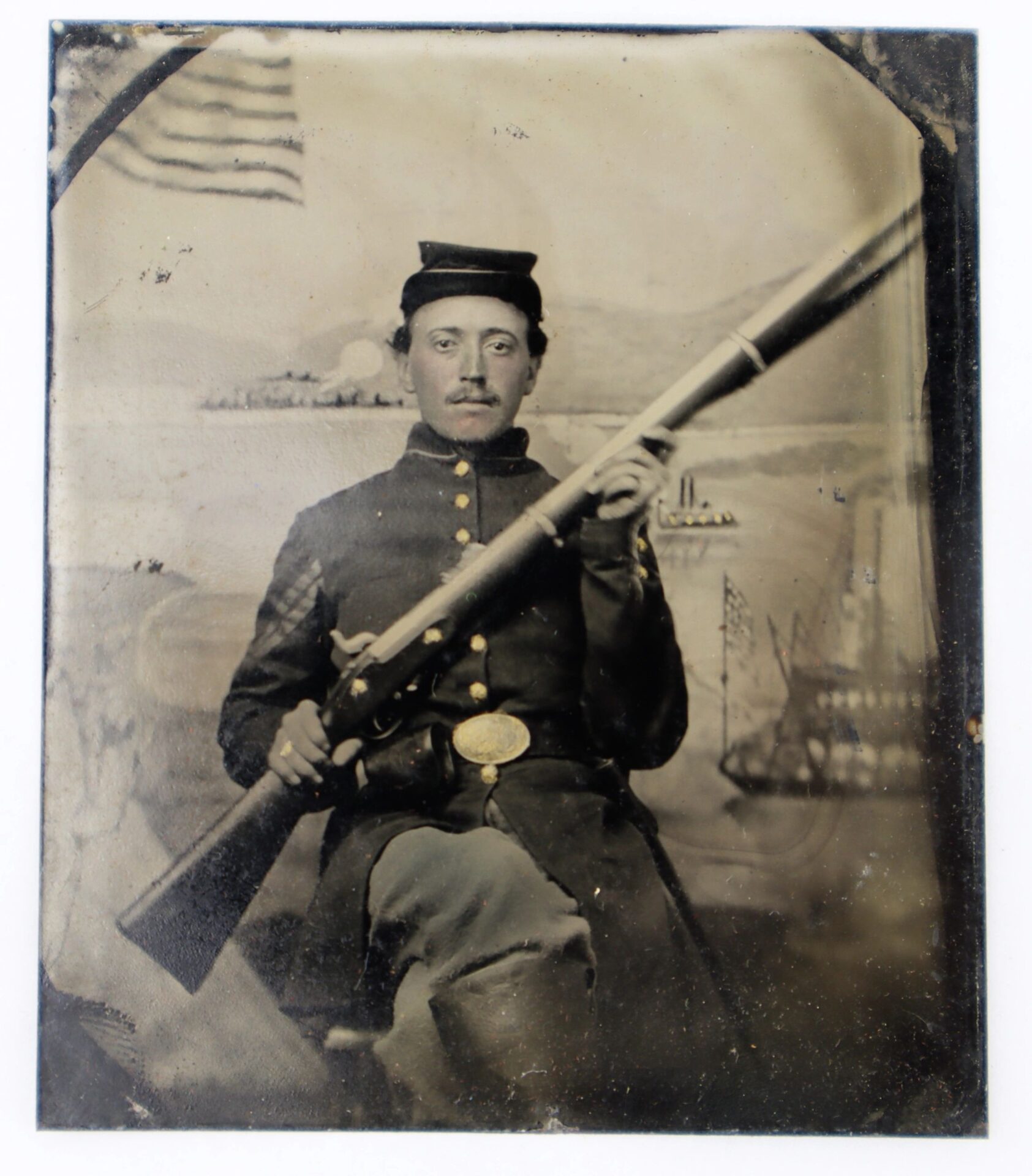 A Union Infantry Corporal, seated and holding an Enfield Rifle Musket, is featured in this 1/6th Plate Tintype, with an American flag and ships visible in the background.