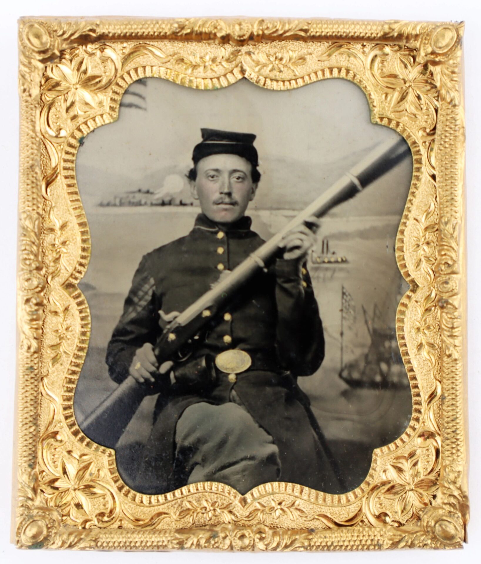 A 1/6th plate tintype of a Union infantry corporal with an Enfield rifle musket, in uniform, framed in an ornate gold case; the painted backdrop shows ships and an American flag.