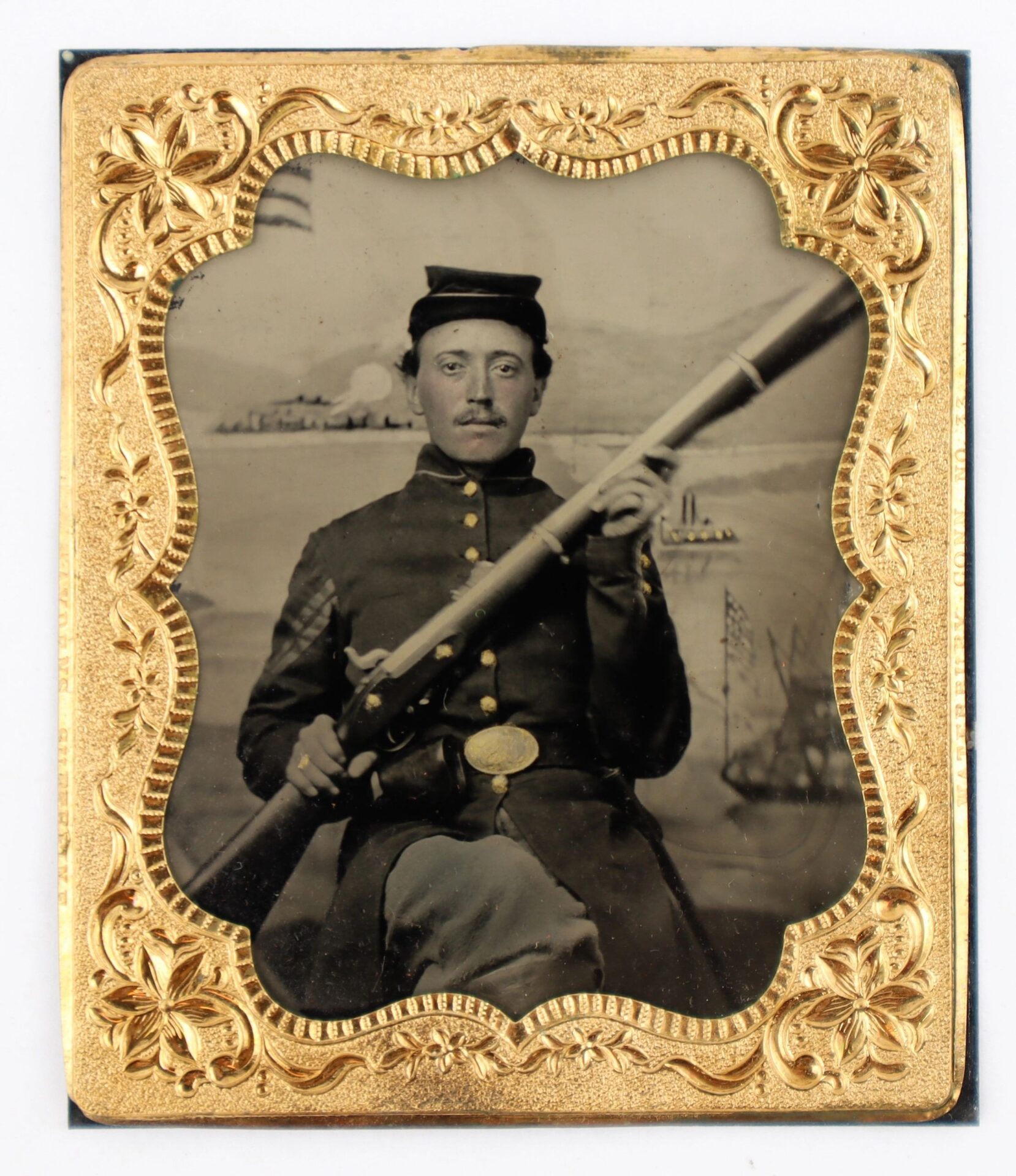 A 1/6th plate tintype shows a Union Infantry Corporal holding an Enfield rifle musket, seated before a painted backdrop of ships and an American flag, framed by an ornate gold border.