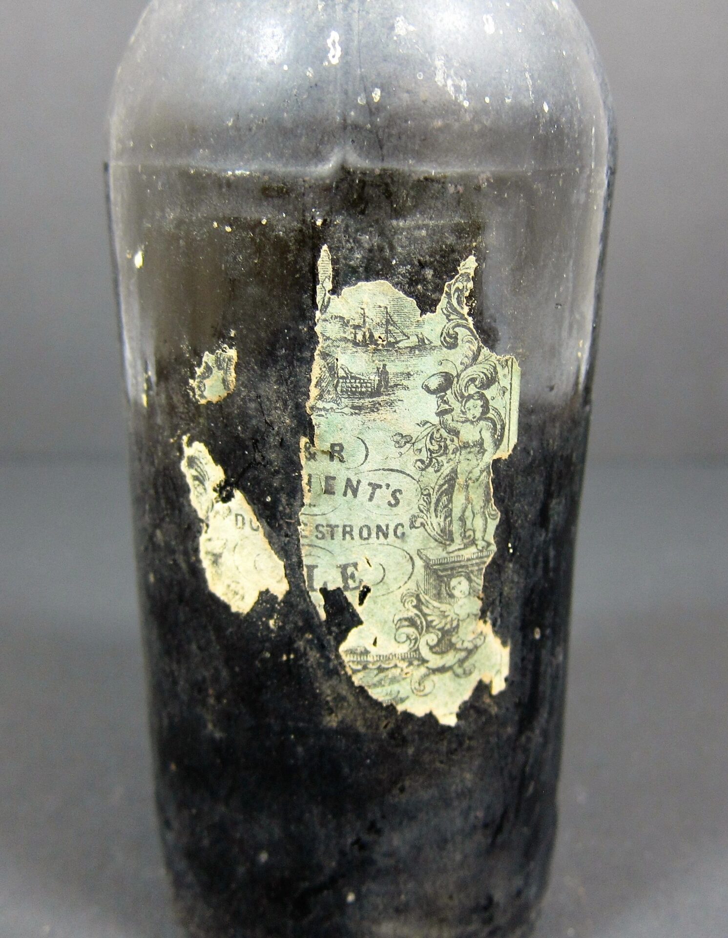 A non-excavated 19th Century Beer Bottle in dark glass, its partially torn, worn label with faded text and images exposes only fragments of the original design.