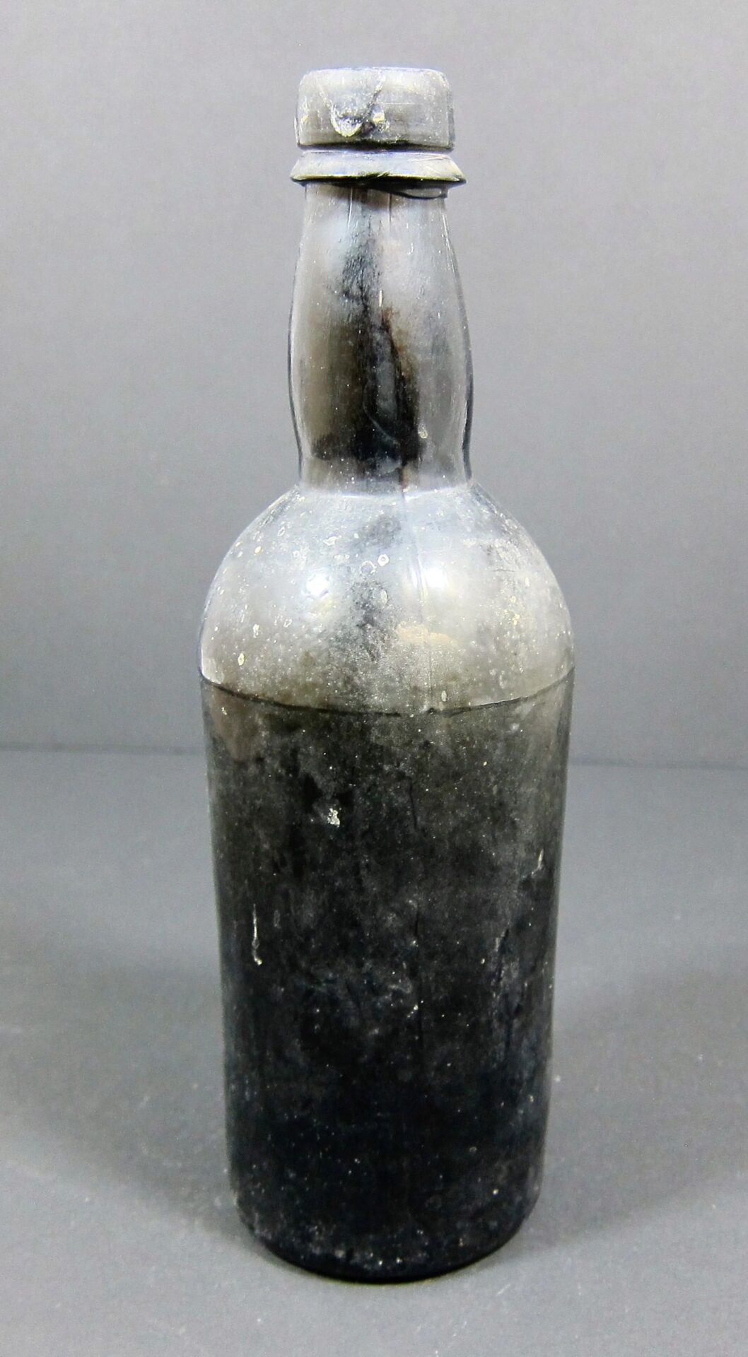 A 19th Century Beer Bottle - Non Excavated, made of dark glass with a cork stopper, stands upright against a plain gray background.