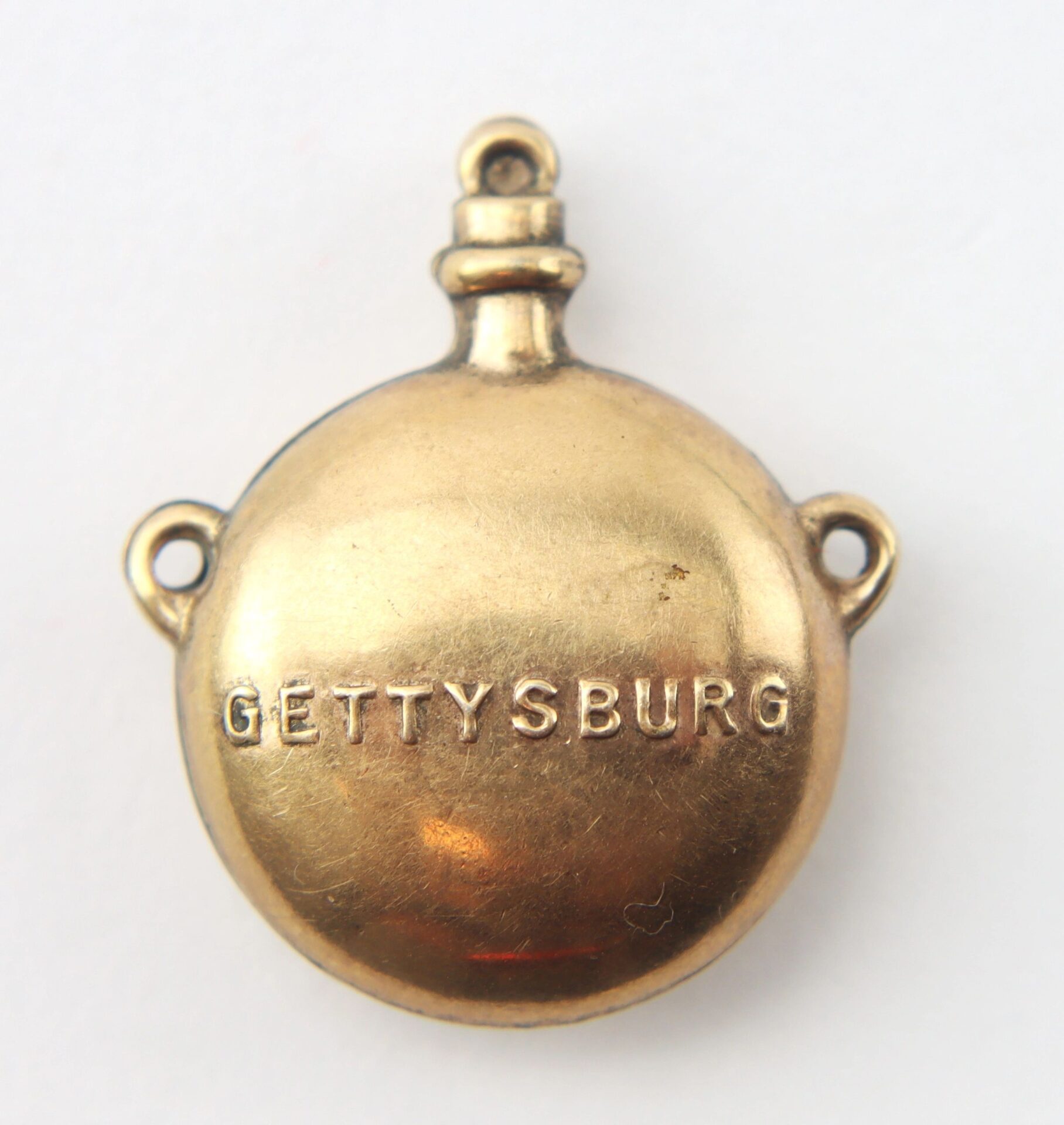 A small, round, gold-toned metal charm shaped like a canteen and engraved with "GETTYSBURG." Product: Gettysburg Souvenir Canteen / SOLD.
