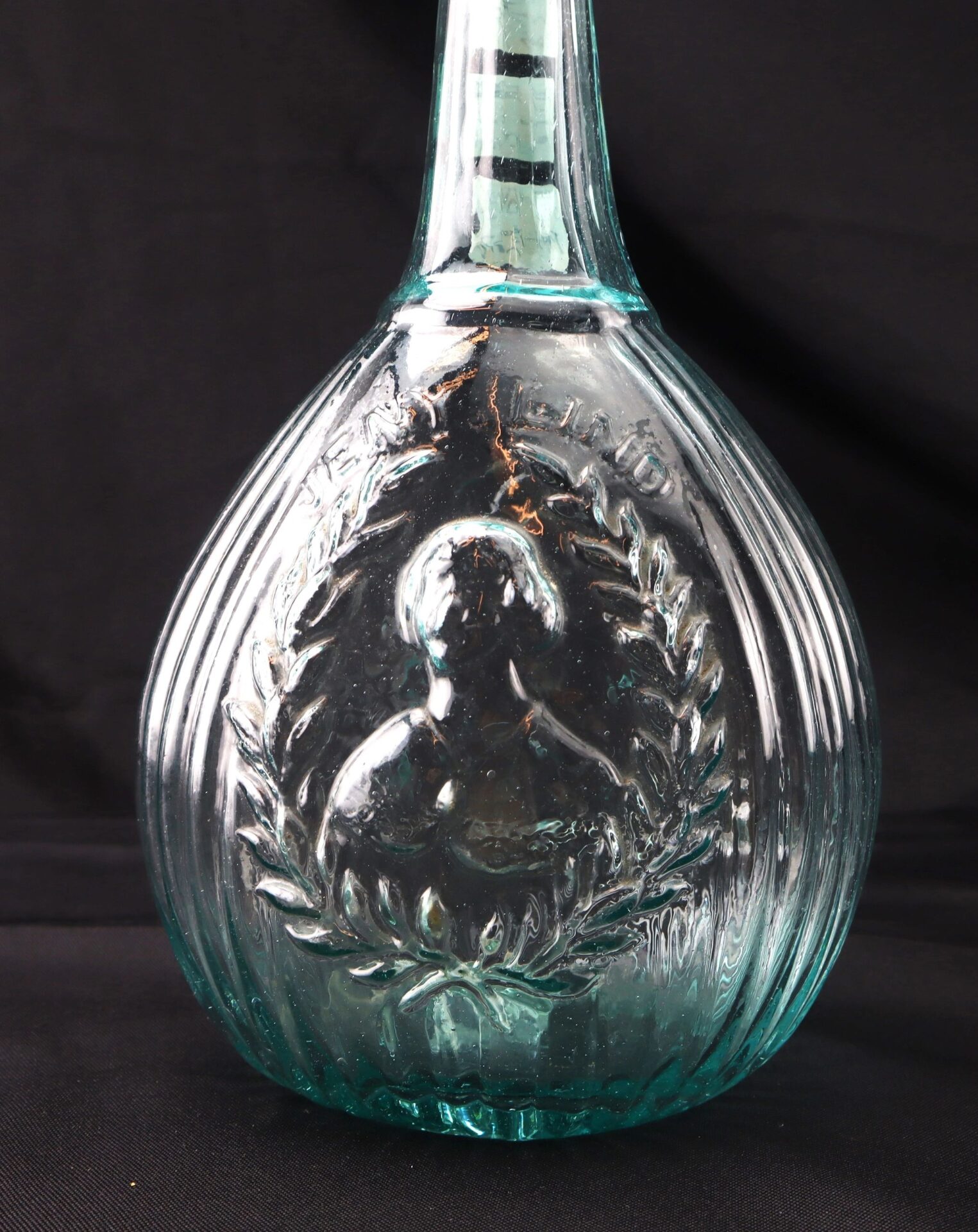 Aqua "Jenny Lind" Flask: Clear glass bottle featuring an embossed portrait of a woman framed by a laurel wreath, displayed against a black background.