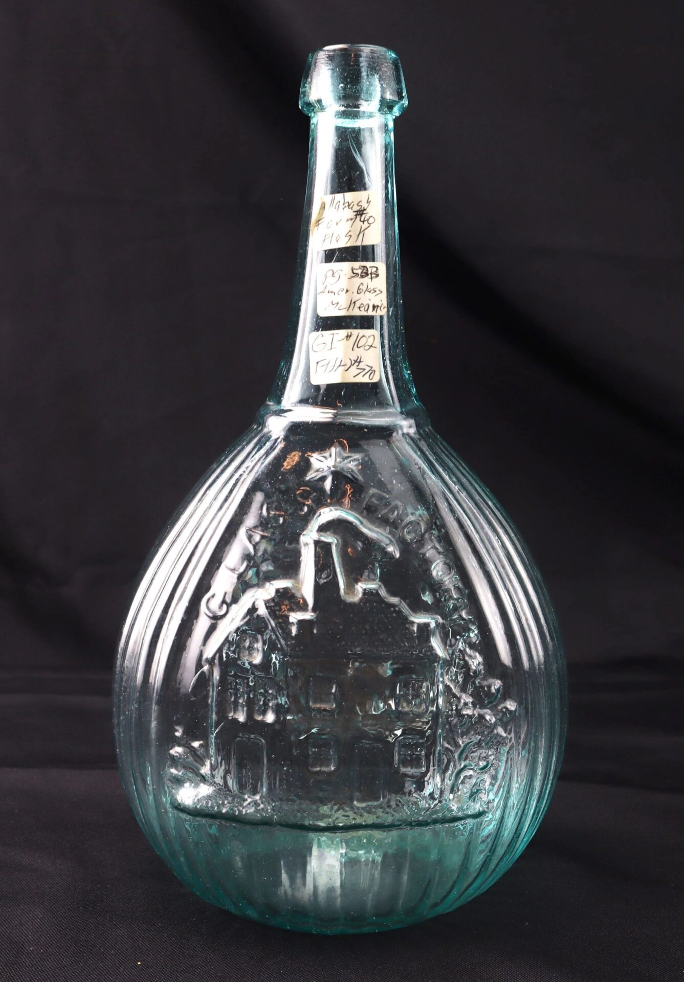 An upright Aqua "Jenny Lind" Flask made of clear embossed glass, featuring a house and tree design, is shown against a black background with handwritten labels attached to its neck.