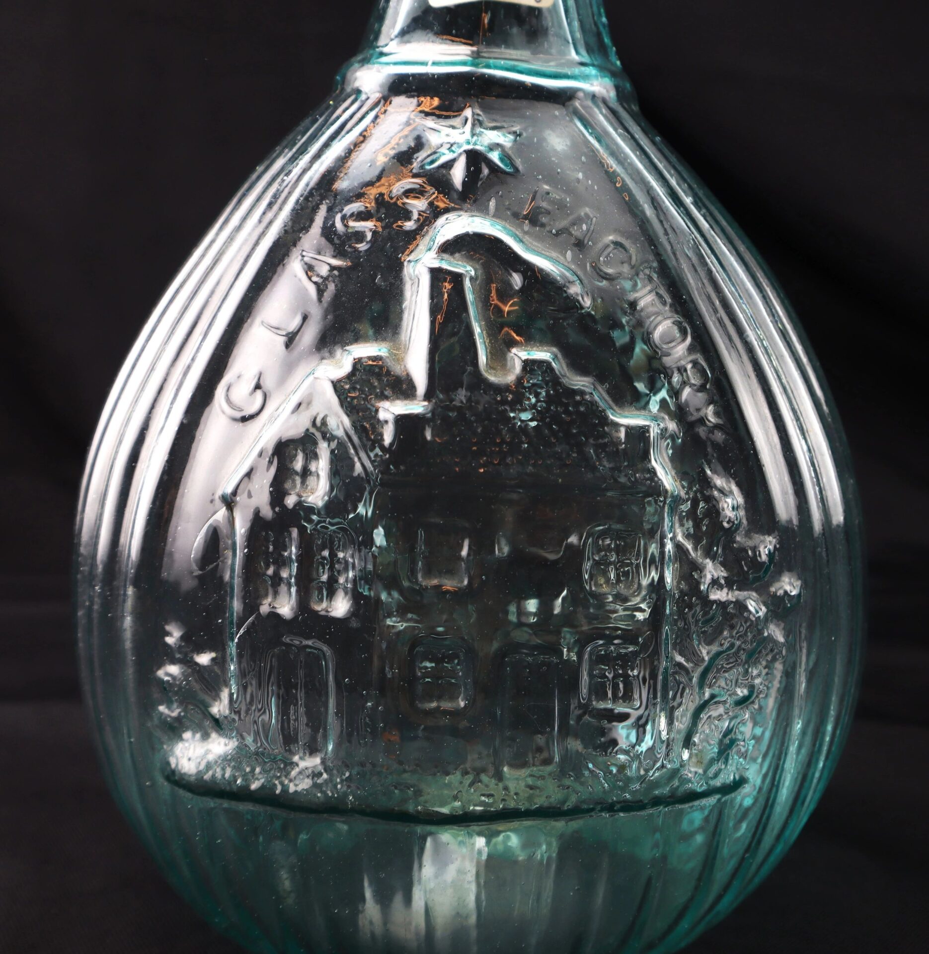 Aqua "Jenny Lind" Flask: teardrop-shaped blue-green glass bottle featuring an embossed factory building and "GLASS FACTORY" on the front, displayed against a black background.