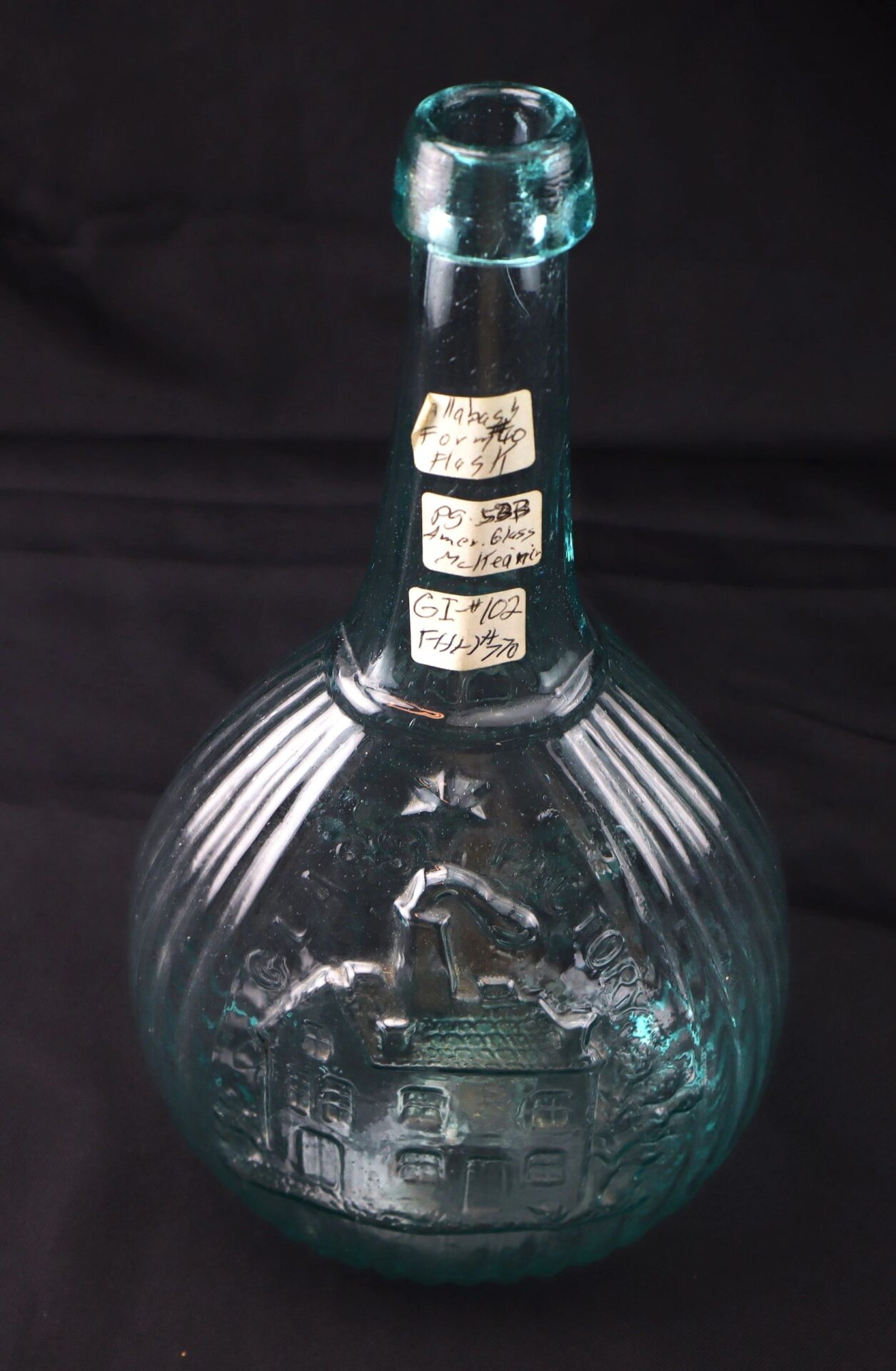 An Aqua "Jenny Lind" Flask with embossed house design and three handwritten neck labels, displayed against a black background.