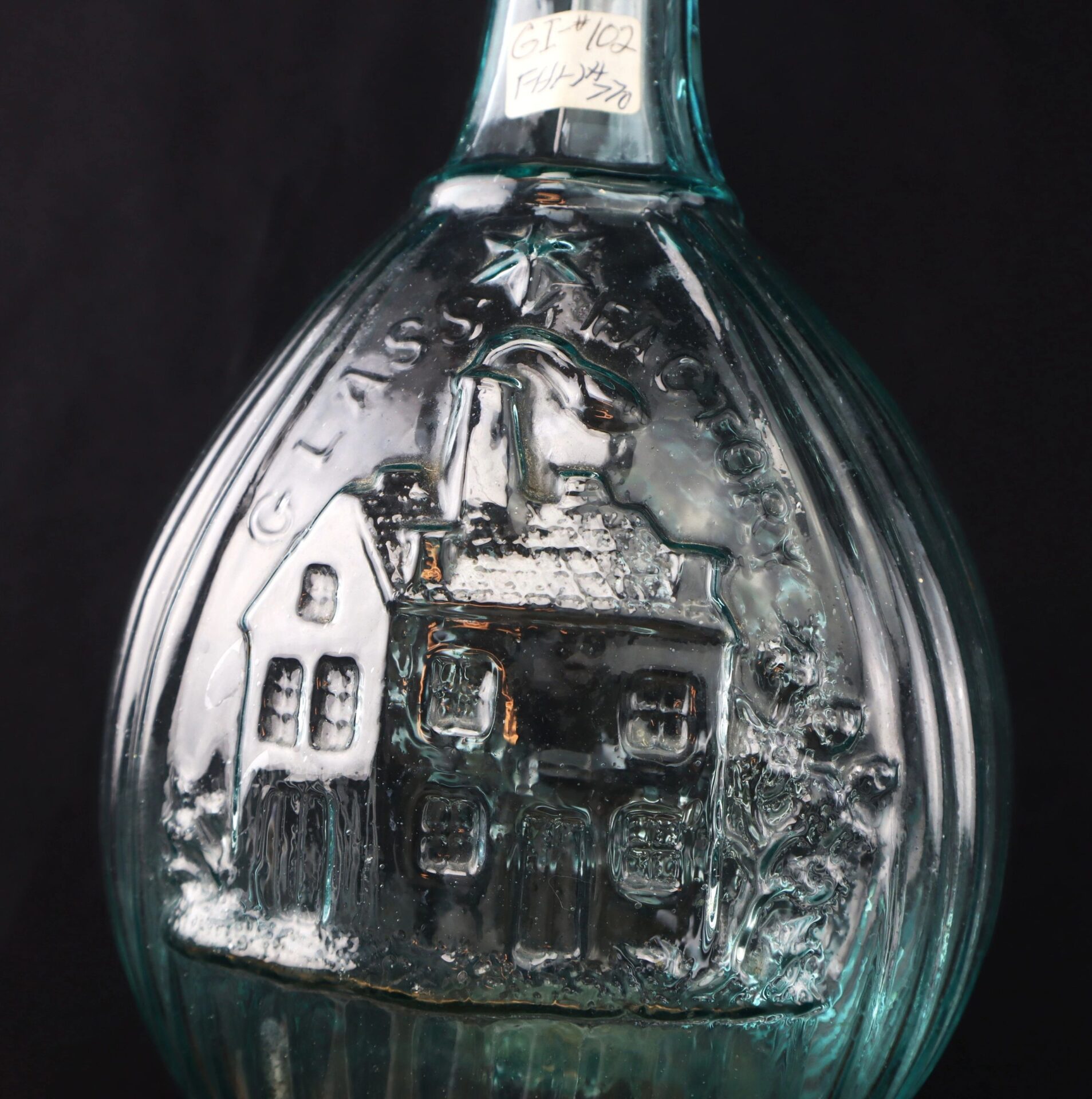 A close-up of an Aqua "Jenny Lind" Flask, a vintage blue-green glass bottle featuring an embossed house and tree design with "GLASS FACTORY" above.
