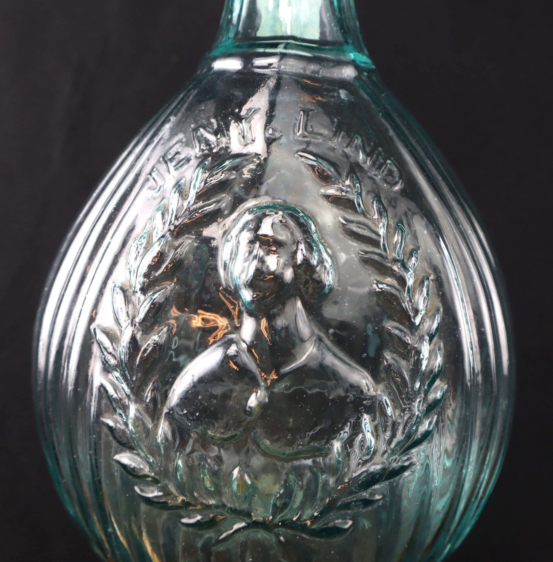 Close-up of the Aqua "Jenny Lind" Flask, an embossed glass bottle featuring a woman's bust framed by laurel leaves and "JENNY LIND" at the top.