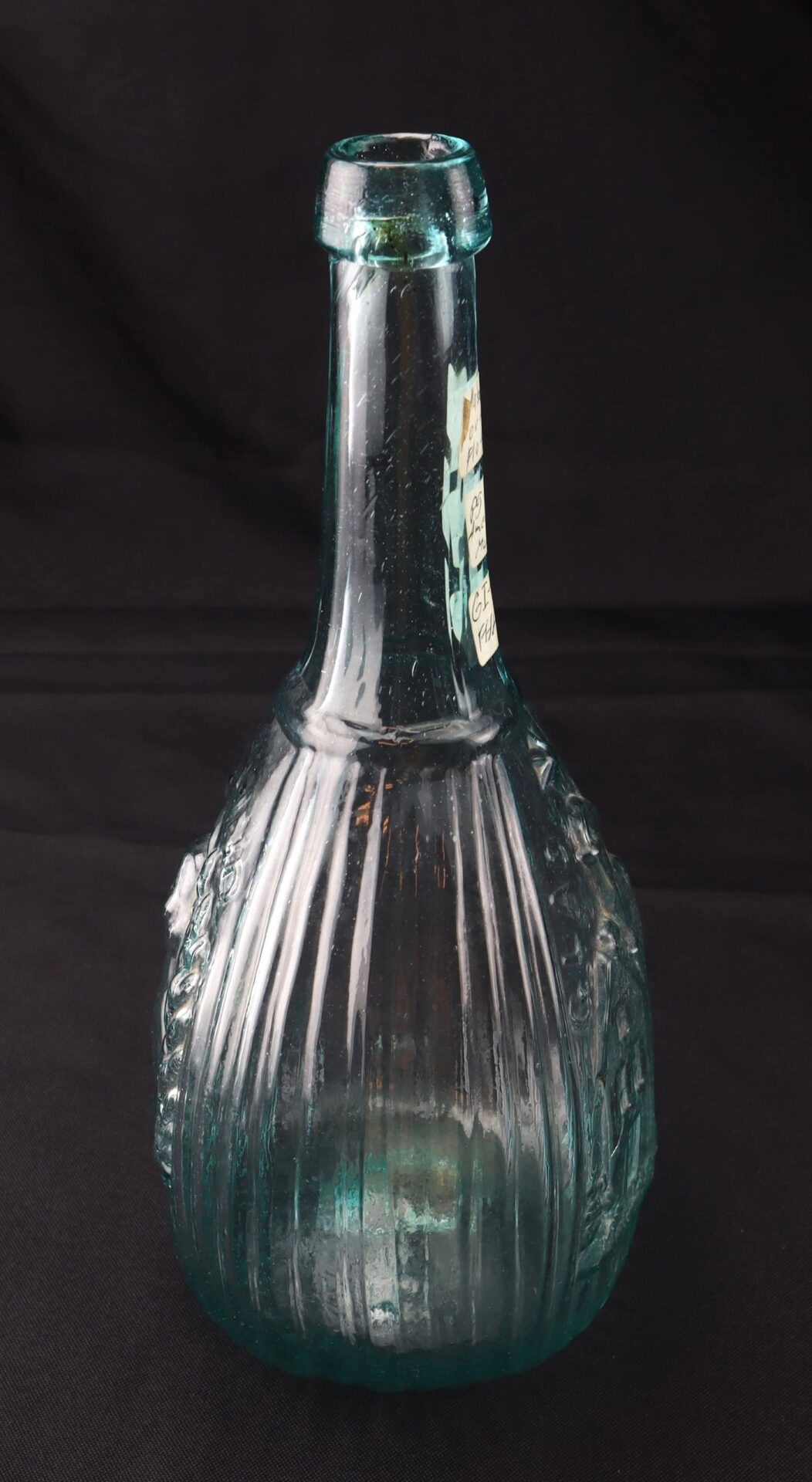 An upright Aqua "Jenny Lind" Flask with a clear, ribbed glass body, long neck, and round base is displayed against a plain dark background.