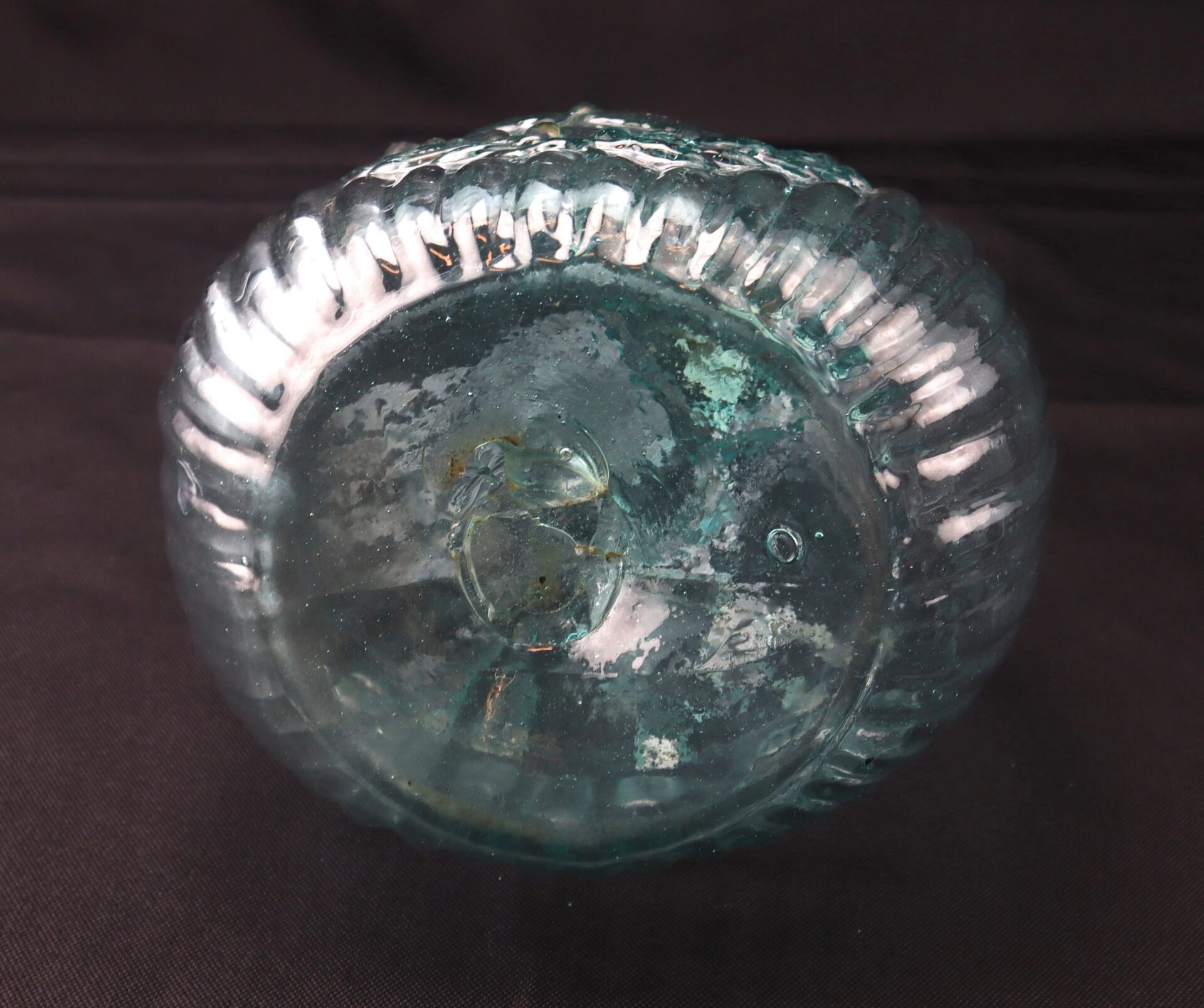 The bottom view of an Aqua "Jenny Lind" Flask, a round ribbed glass bottle with clear blue-green color and visible imperfections, shown against a dark background.