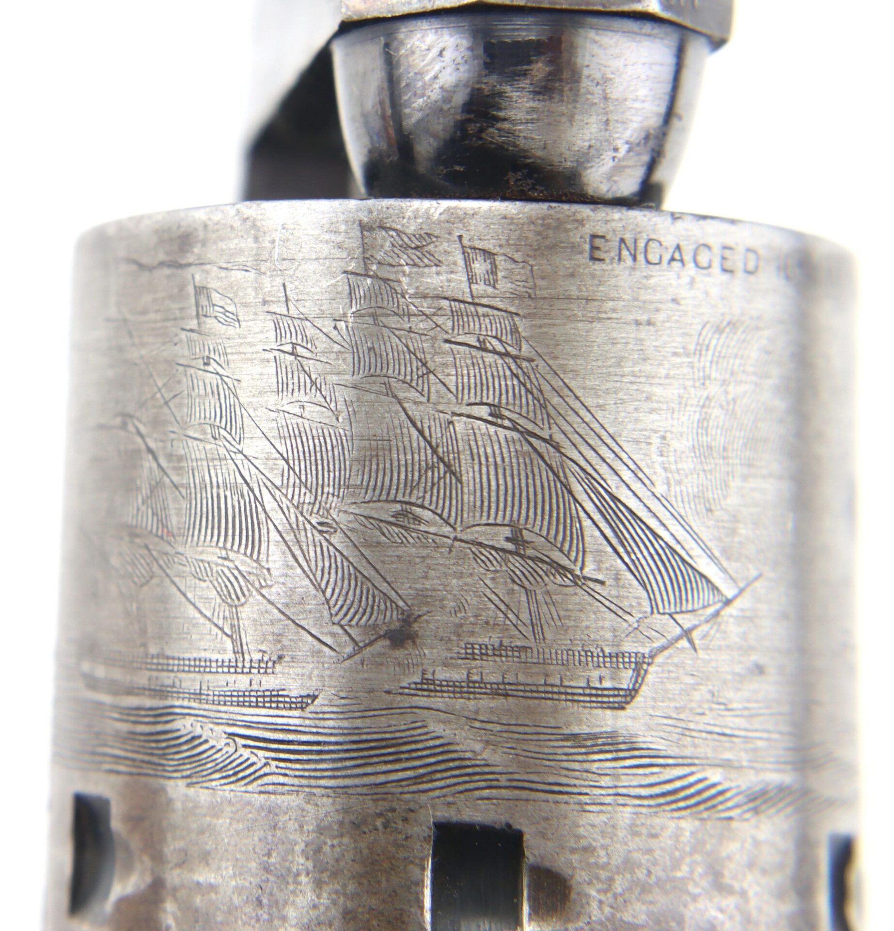 Close-up of a metal cylinder from a Cased London Colt 1851 Navy Percussion Revolver, engraved with sailing ships and the word "ENGAGED" partially visible at the top right.
