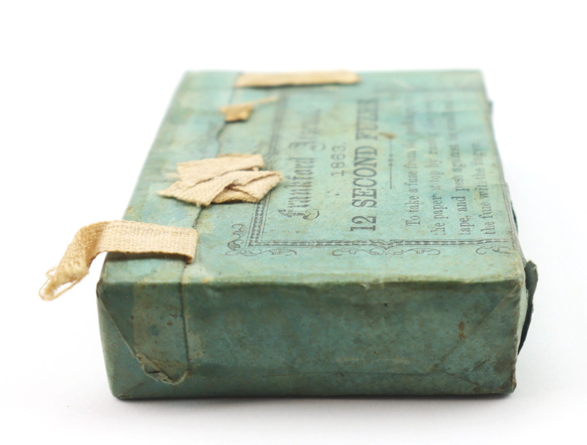 A rectangular, vintage blue-green box labeled "Frankford Arsenal 12 Second Fuses" features cream fabric ties and faded text on the lid.