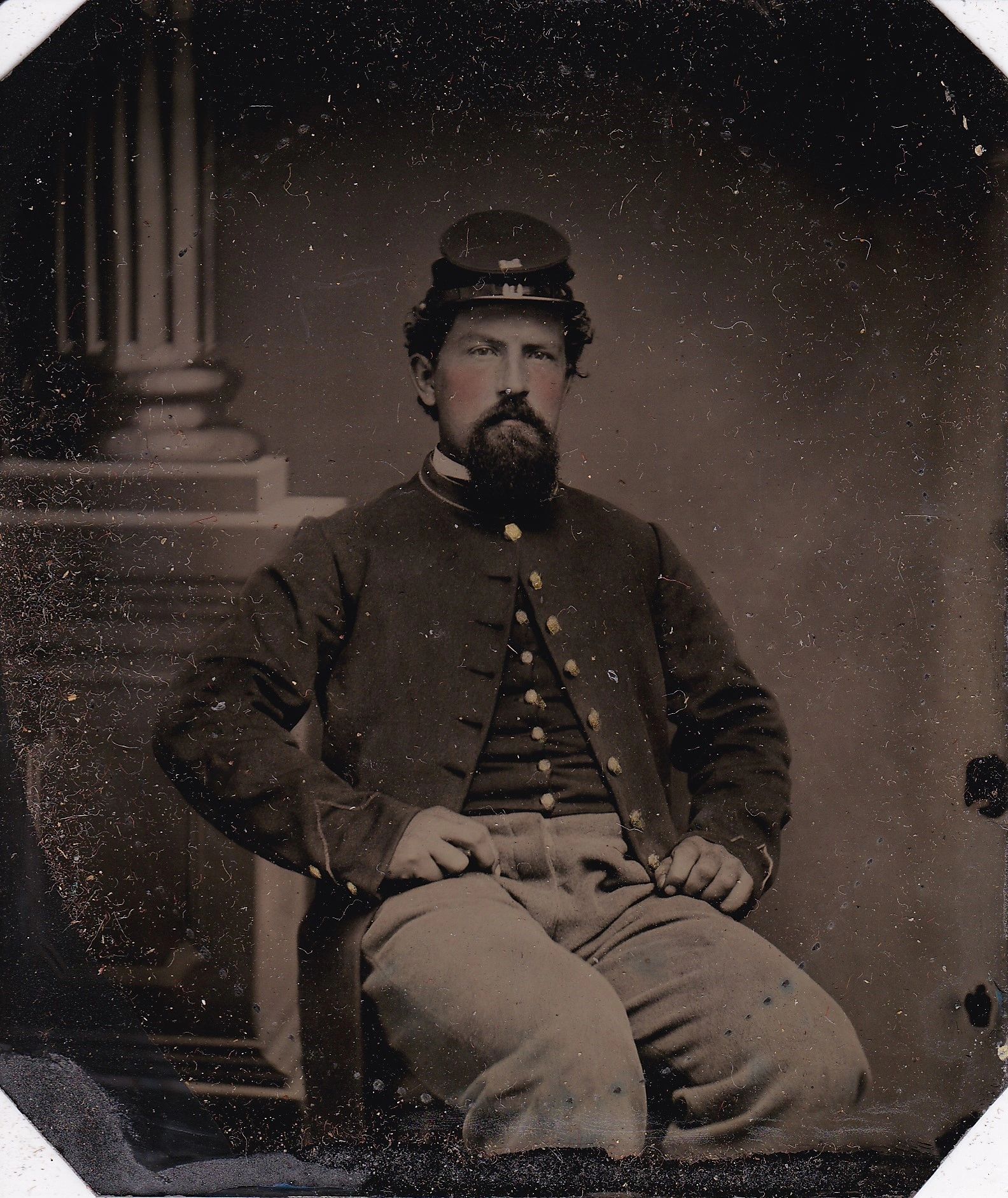 A bearded man in a dark jacket and cap sits on a chair with his hands on his hips; a column is in the background. This scene appears in the Union Engineer Sixth Plate Tintype.