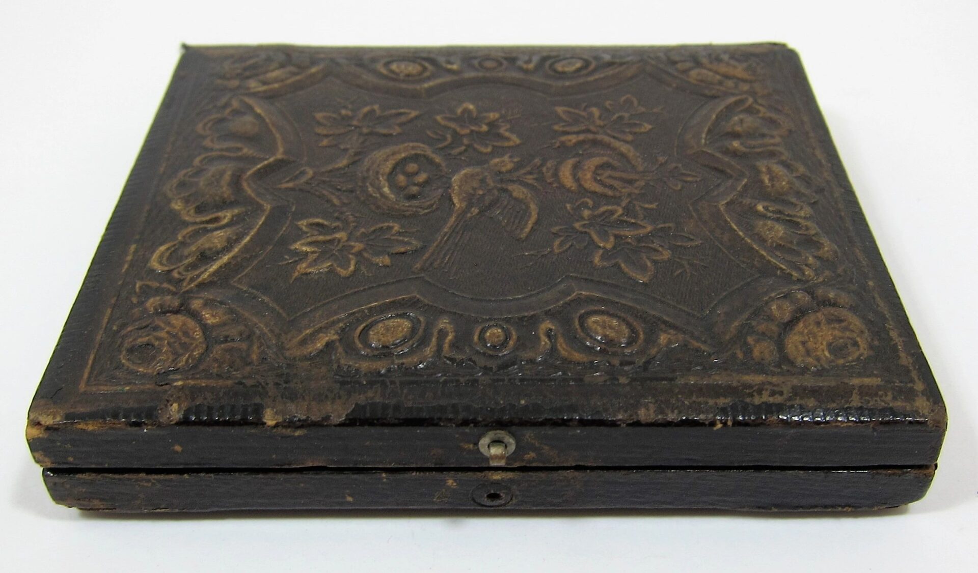A closed, old, dark brown leather case with floral and bird embossing and edge wear containing a Union Engineer Sixth Plate Tintype.