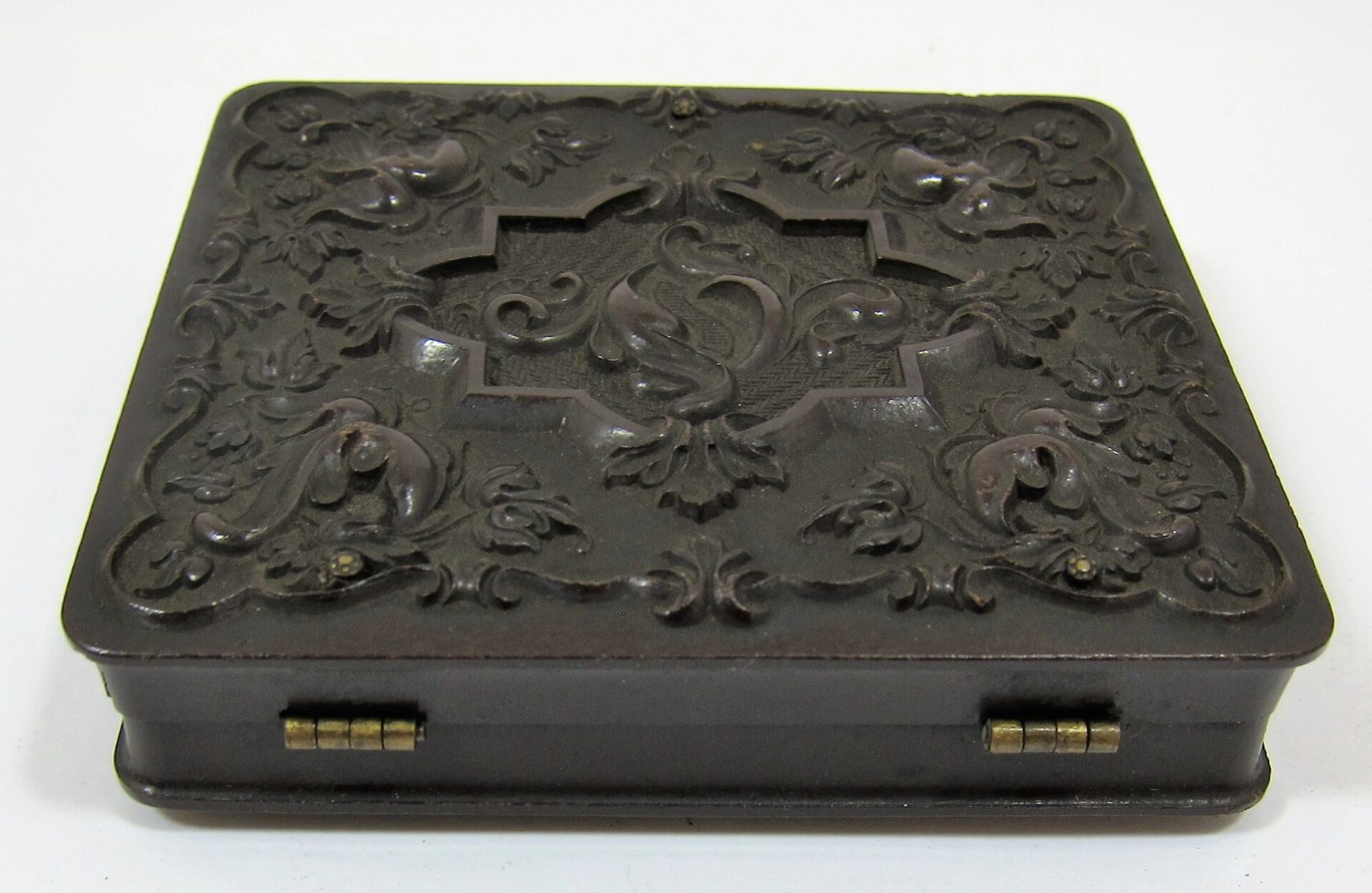 A closed, square, dark brown case with ornate floral and scrollwork designs on the lid and small brass hinges along the front edge houses the Infantry Officer Holding Musket Sixth Plate Tintype.