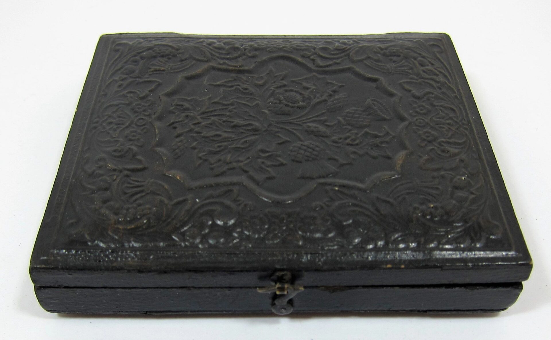 The Soldier with Model 1816 Musket Sixth Plate Tintype is displayed in a closed, ornate black case with embossed floral patterns and a small metal clasp, set against a plain white background.