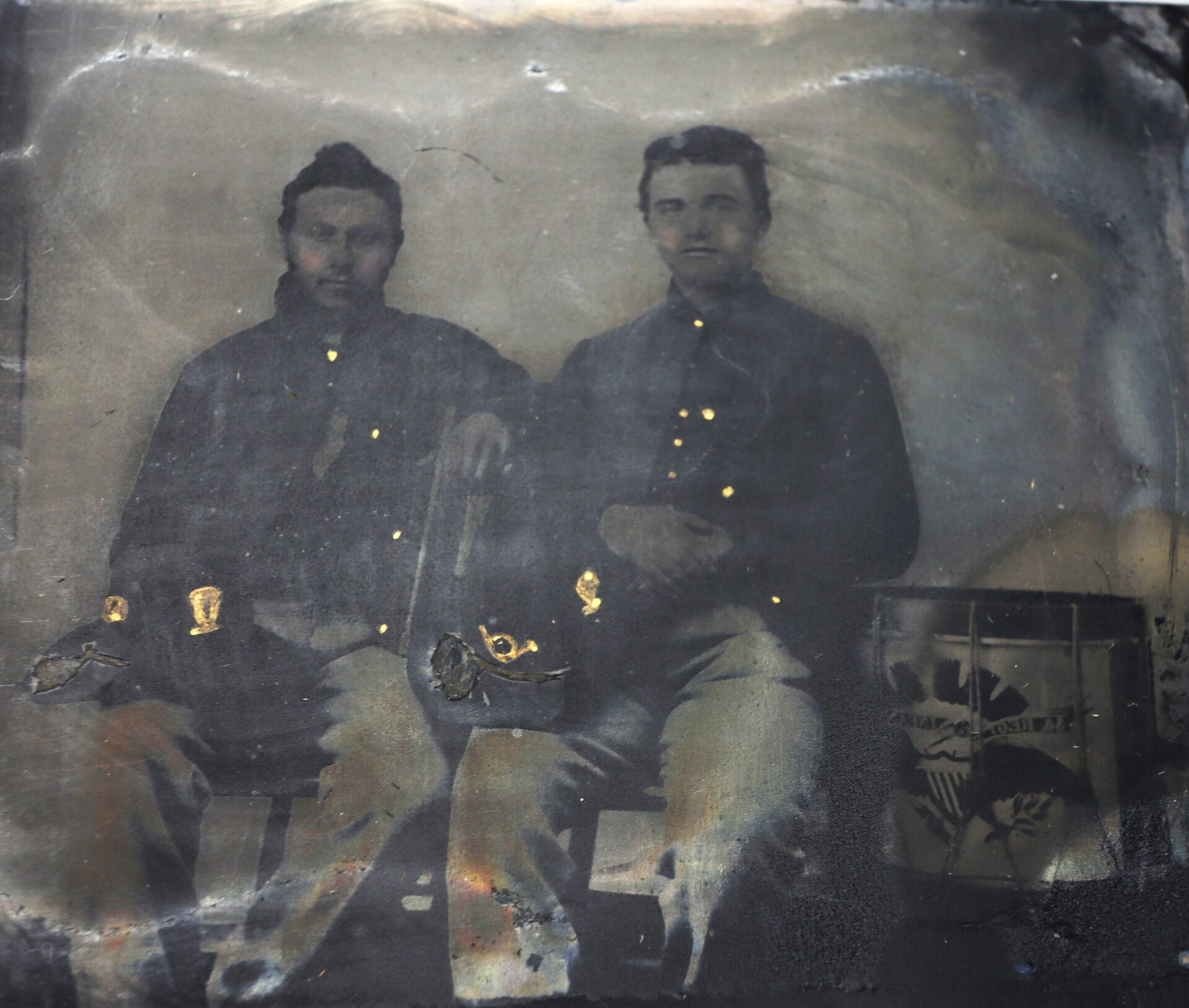In this 1/4 Plate Tintype, two Union musicians in Hardee hats sit side by side on a bench next to a drum with an eagle design in this faded, antique photograph.