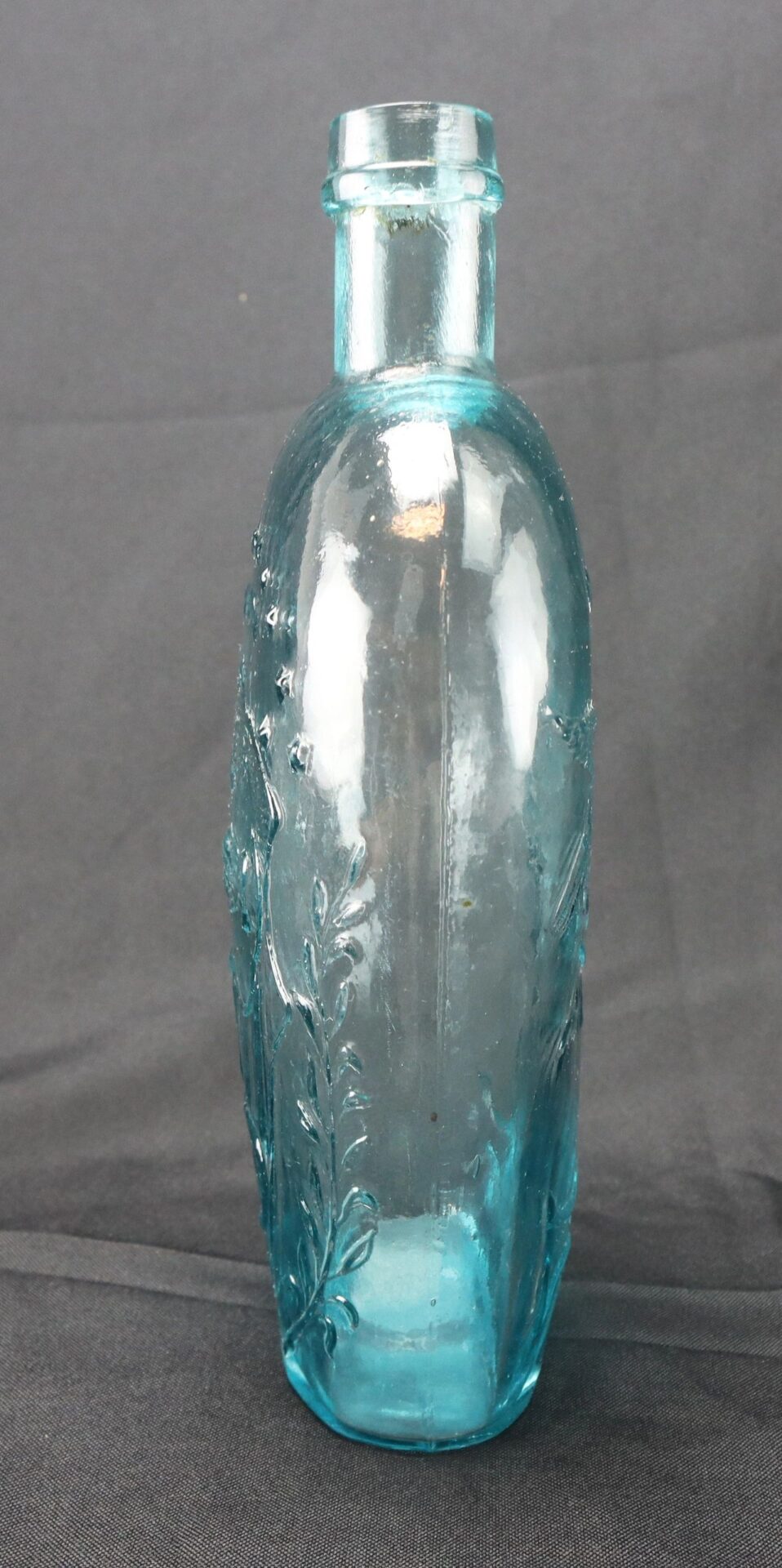 A Civil War-Era Union Patriotic Flask made of aqua blue glass with embossed leaf designs and a narrow neck, shown standing upright on a dark surface.