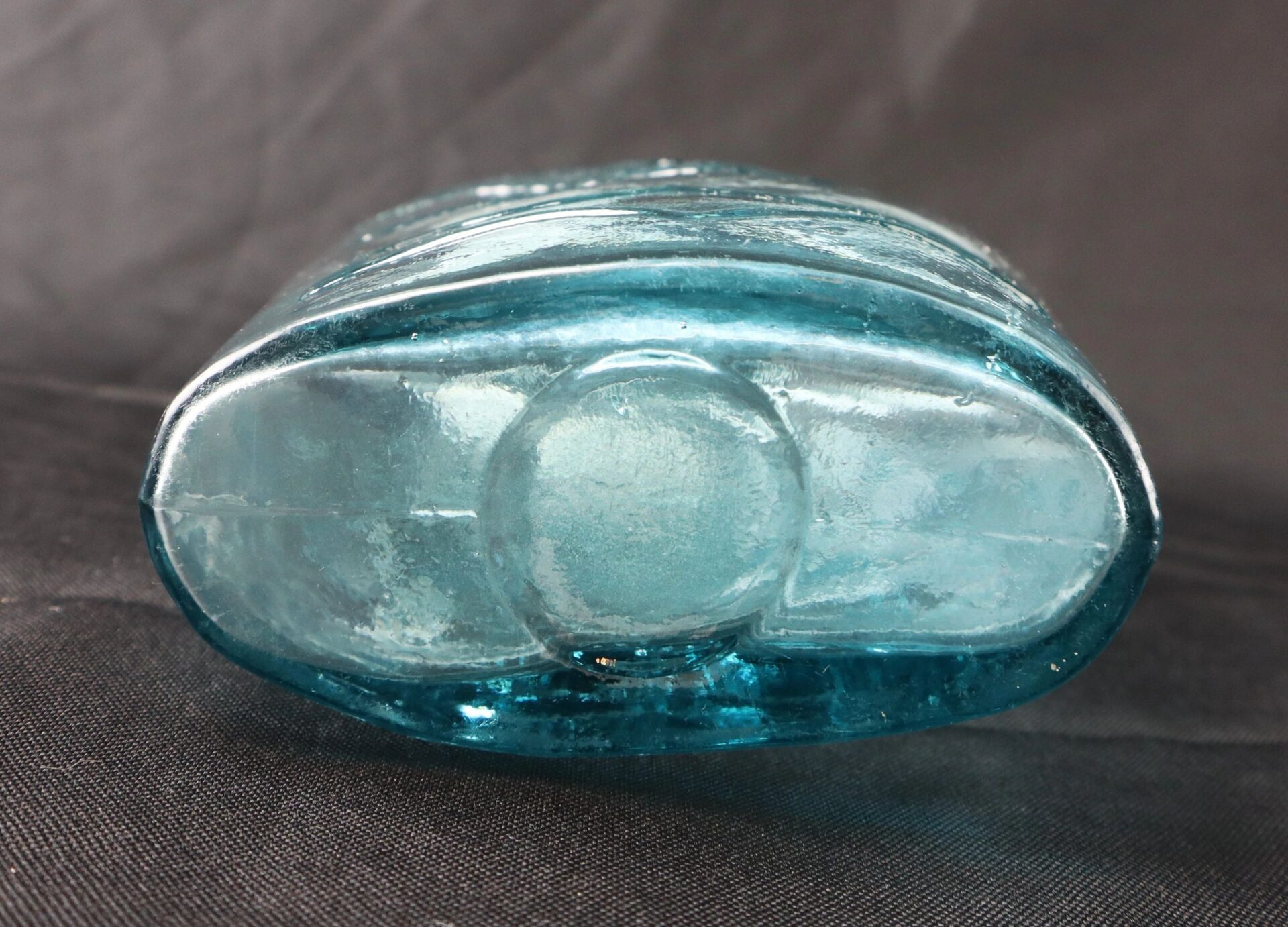 A close-up of the bottom of the Civil War-Era Union Patriotic Flask in aqua blue glass, featuring a central circular indentation, displayed on a dark surface.