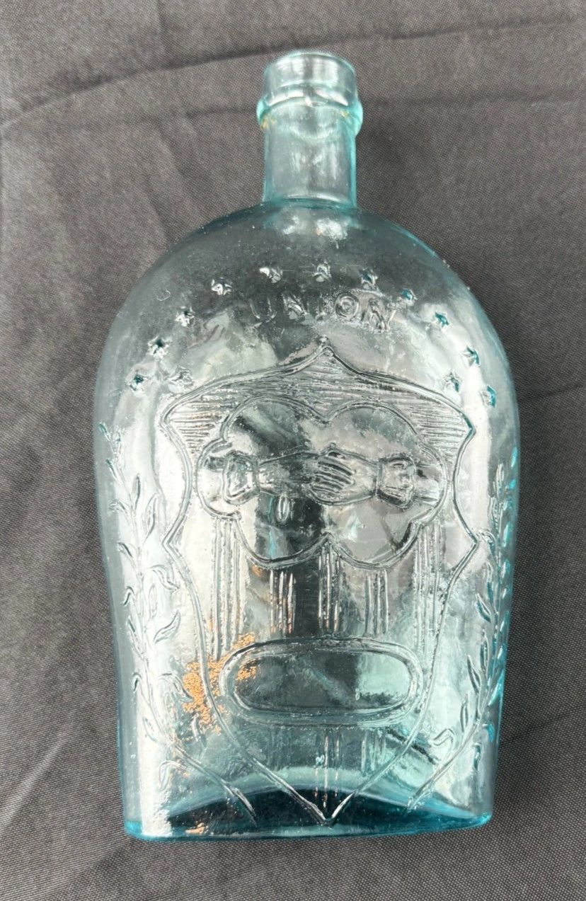 A Civil War-Era Union Patriotic Flask in aqua blue glass, featuring an embossed design of clasped hands with stars and leaves, displayed on a dark fabric background.