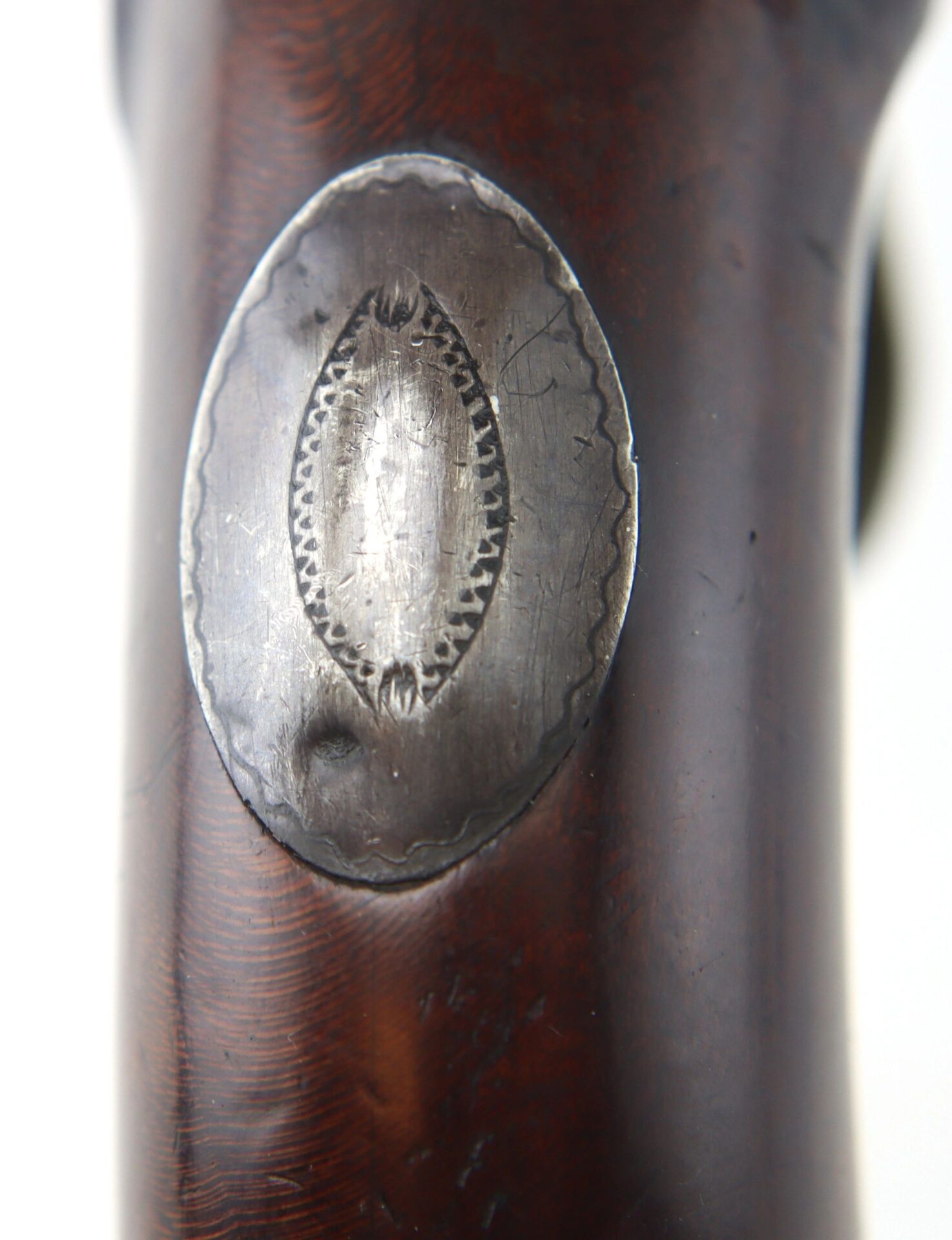 Close-up of an English pistol with a brass barrel and grotesque mask butt cap, featuring a metal oval plate with decorative edges mounted on wood, displaying signs of wear and scratches.