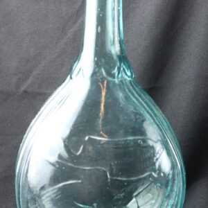 A clear, greenish Historic Glass Flask featuring clasped hands and an eagle motif, with a rounded base and long narrow neck, set against a black background.