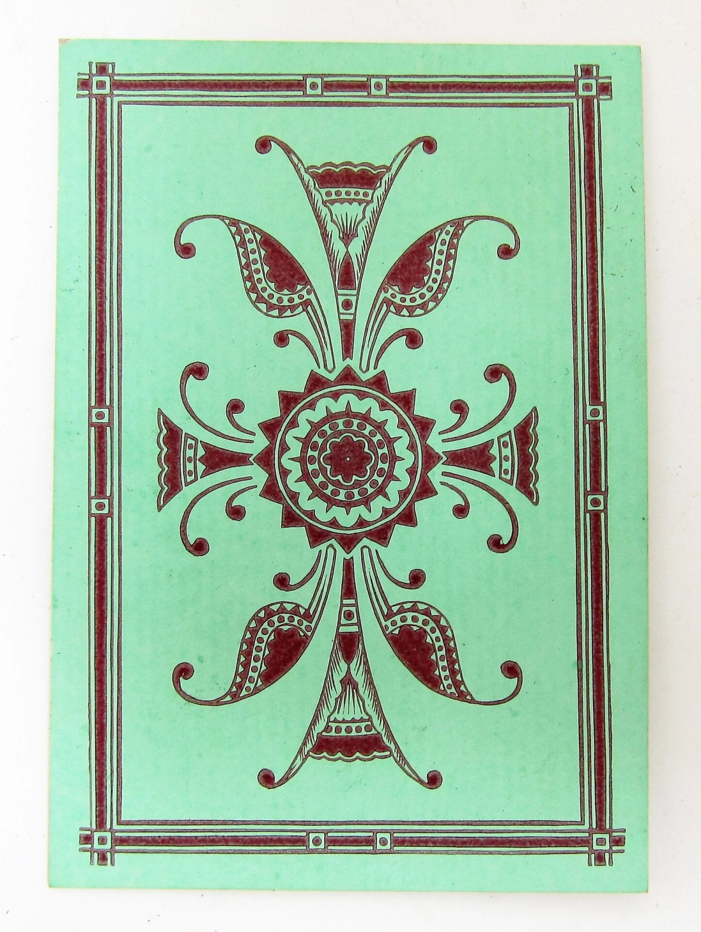 Bezique Card Game features a green card with a symmetrical dark red floral pattern, double-line border, and small square accents in each corner.