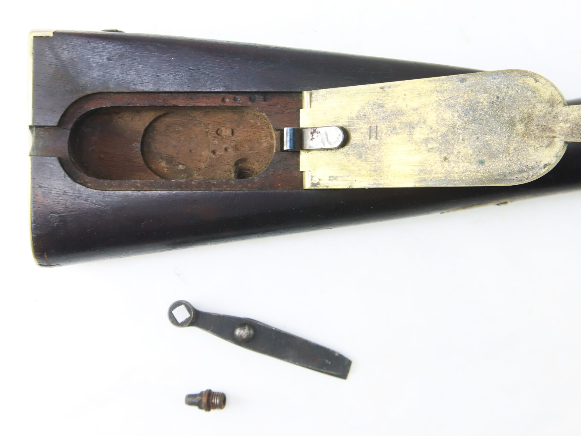 Close-up of a rifle buttstock with the brass buttplate removed, exposing an internal compartment and disassembled metal parts laid nearby.