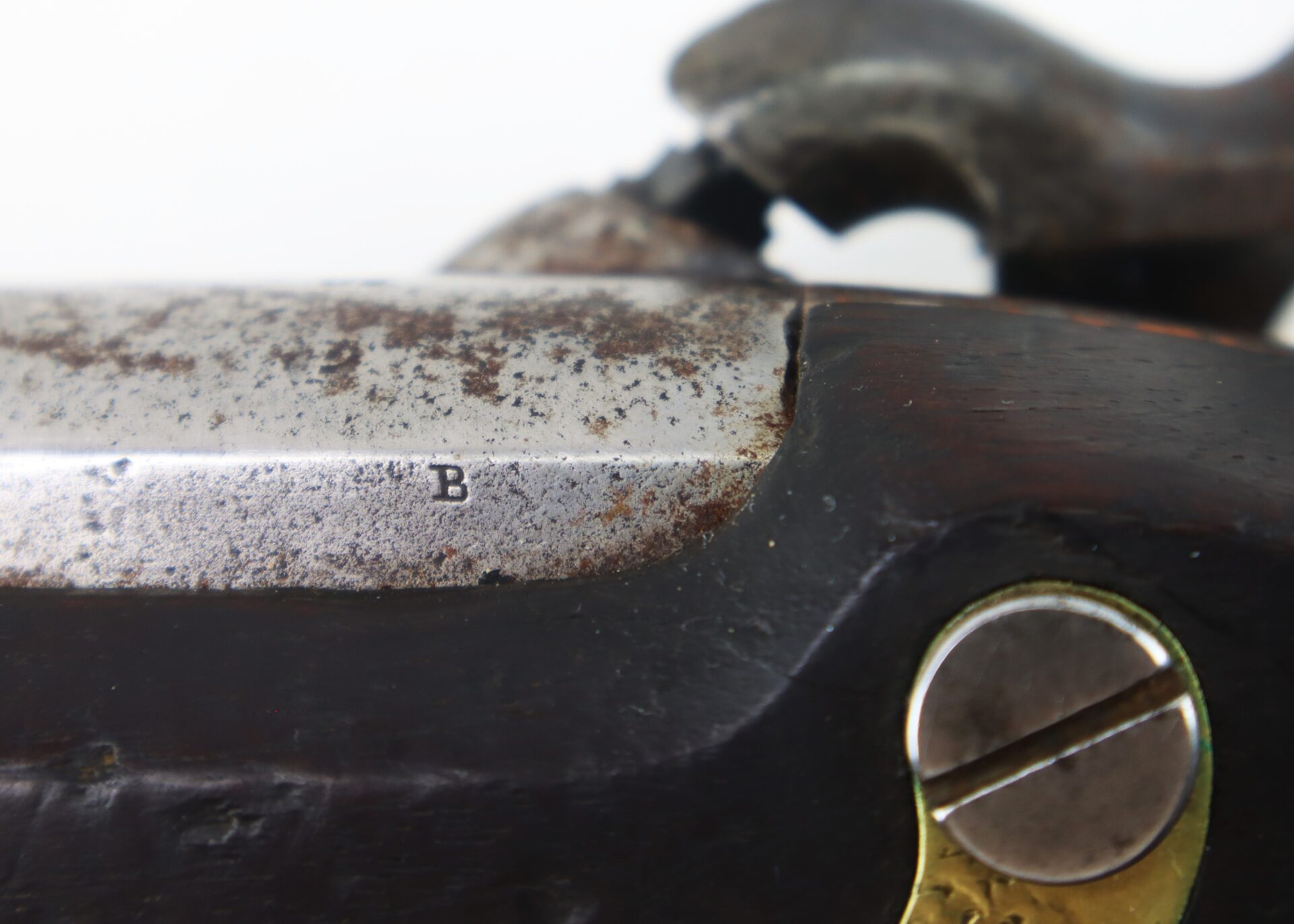 Close-up of an old, weathered firearm showing a metal part with a "B" mark, wood grip, and a brass screw.