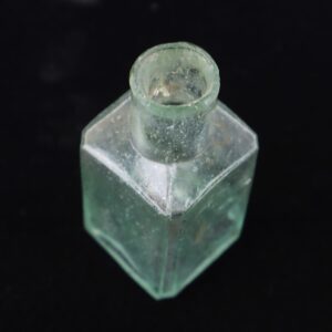 A Civil War Ink Well: a small, clear glass bottle with a square base and short round neck, set against a black background.